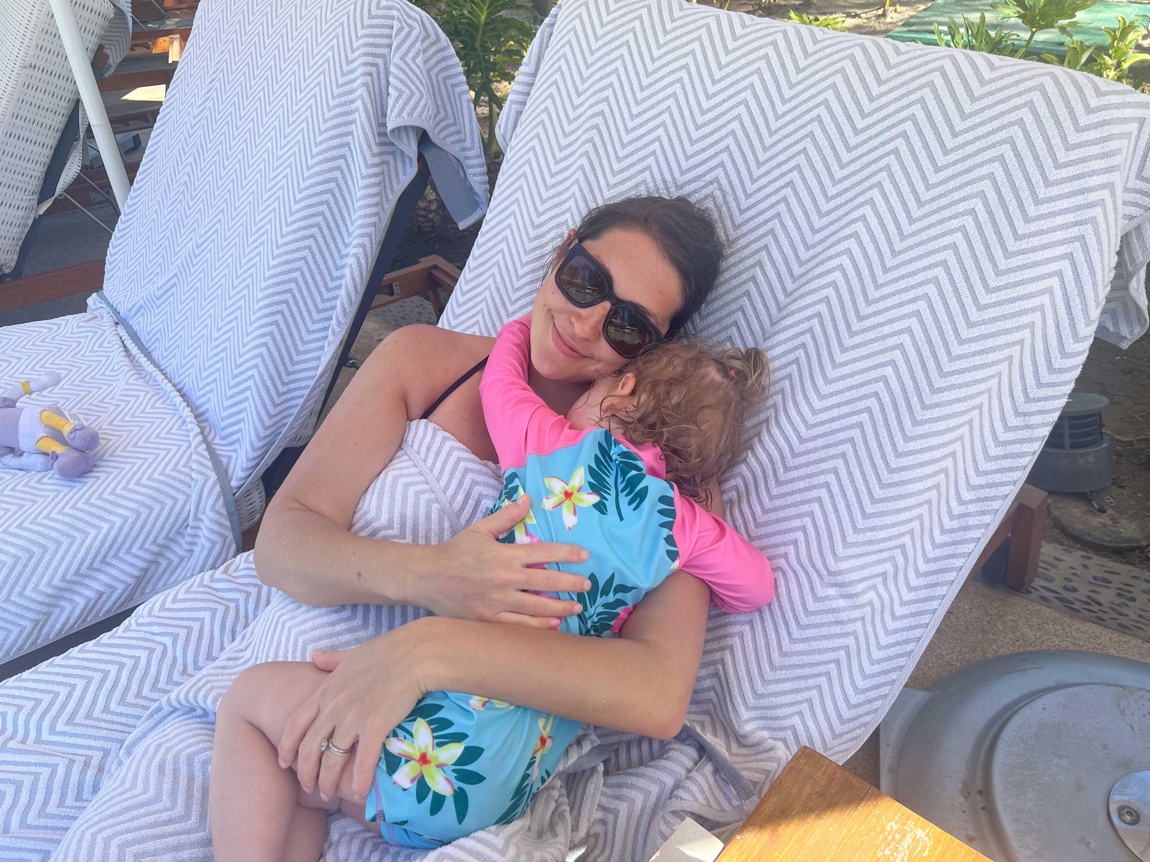 Eine Frau mit Sonnenbrille umarmt ihr Kind auf einem mit Handtüchern bedeckten Liegestuhl.