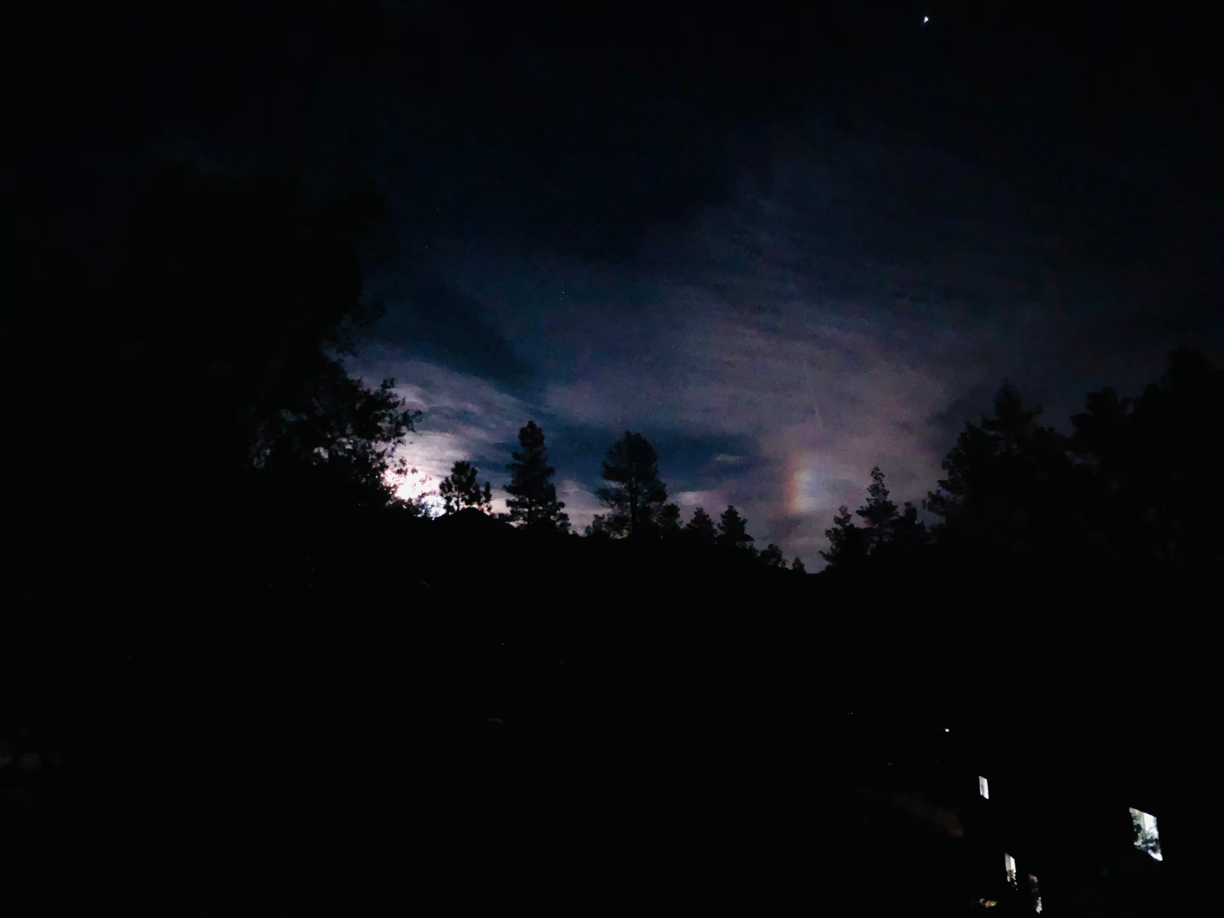Ein Bild eines „Mondbogens“, eines Regenbogens, der aus dem Mond entsteht, nachts durch die Bäume und Wolken.