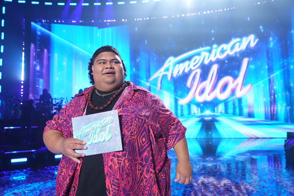 Der hawaiianische Künstler Iam Tongi steht auf dem leuchtend blauen Set von American Idol, mit dem Namen der Show im Hintergrund und einem Lächeln im Gesicht, auf einem undatierten Bild.