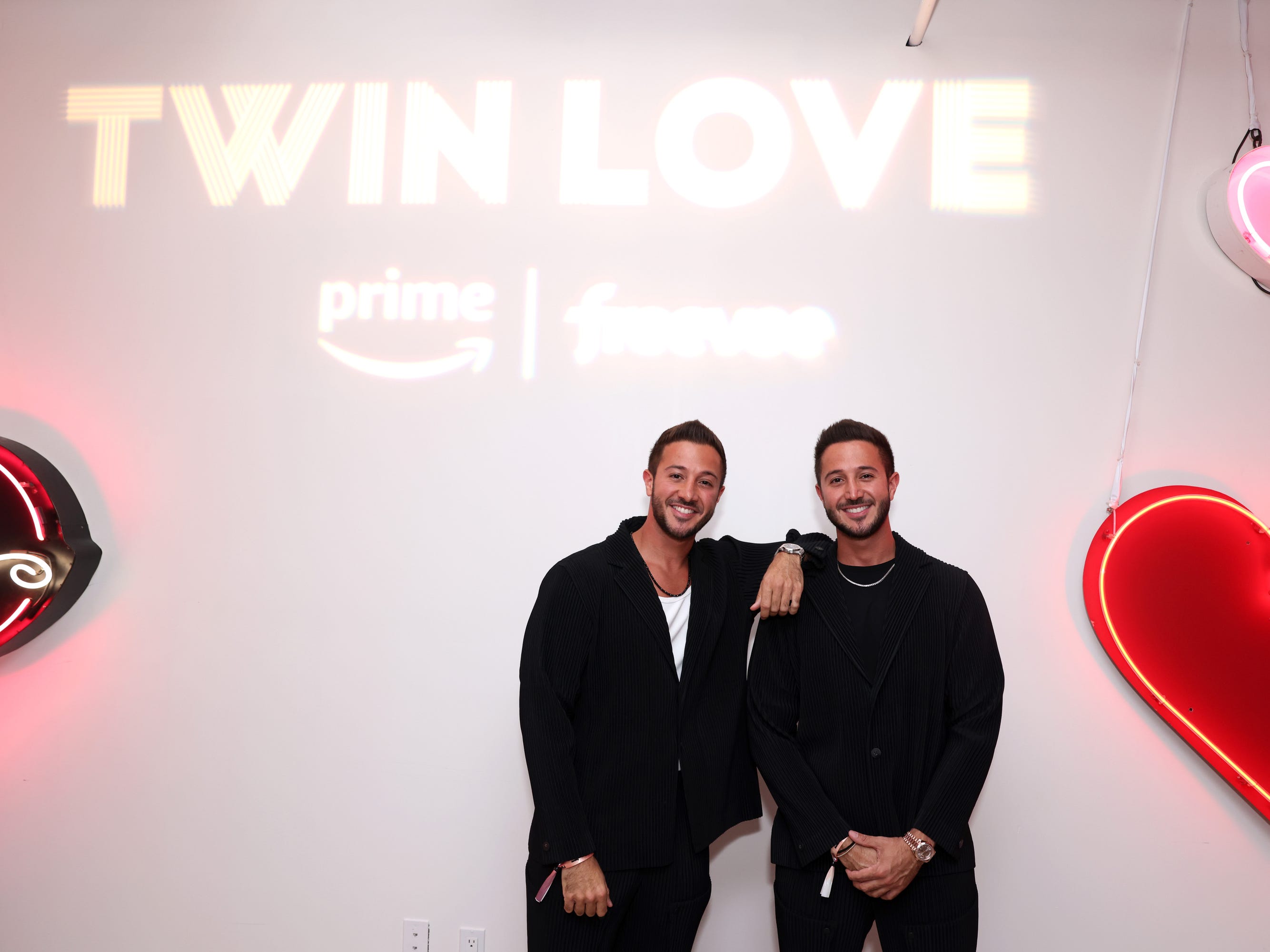 Samer und Samir Akel in schwarzer Kleidung, lächelnd unter einem Schild mit der Aufschrift „Zwillingsliebe“.