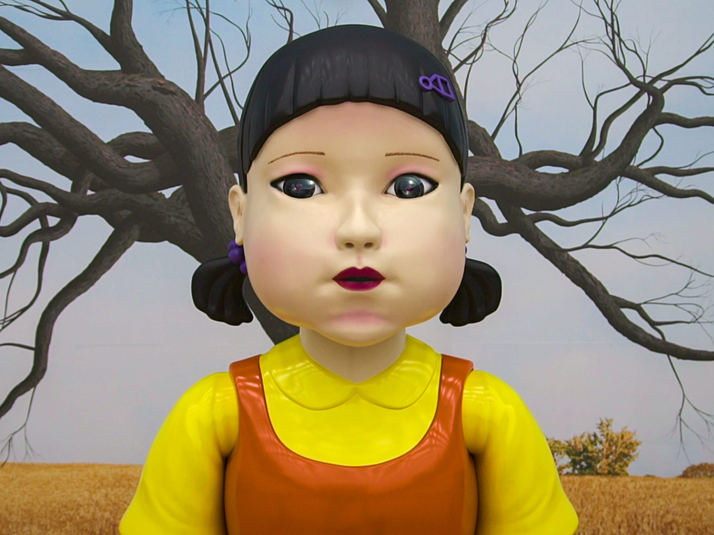 Die große Puppe im Tintenfisch-Spiel stellt die Herausforderung dar, eine kindliche Figur mit gelbem Hemd, orangefarbenem Kleid und Zöpfen