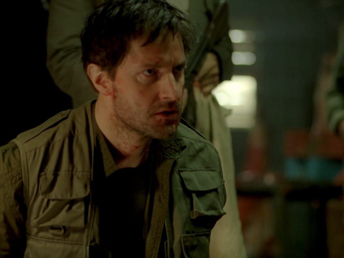 Richard Armitage als John Porter, der mit gefesselten Händen auf dem Boden kauert, während jemand eine Waffe hinter sich hält
