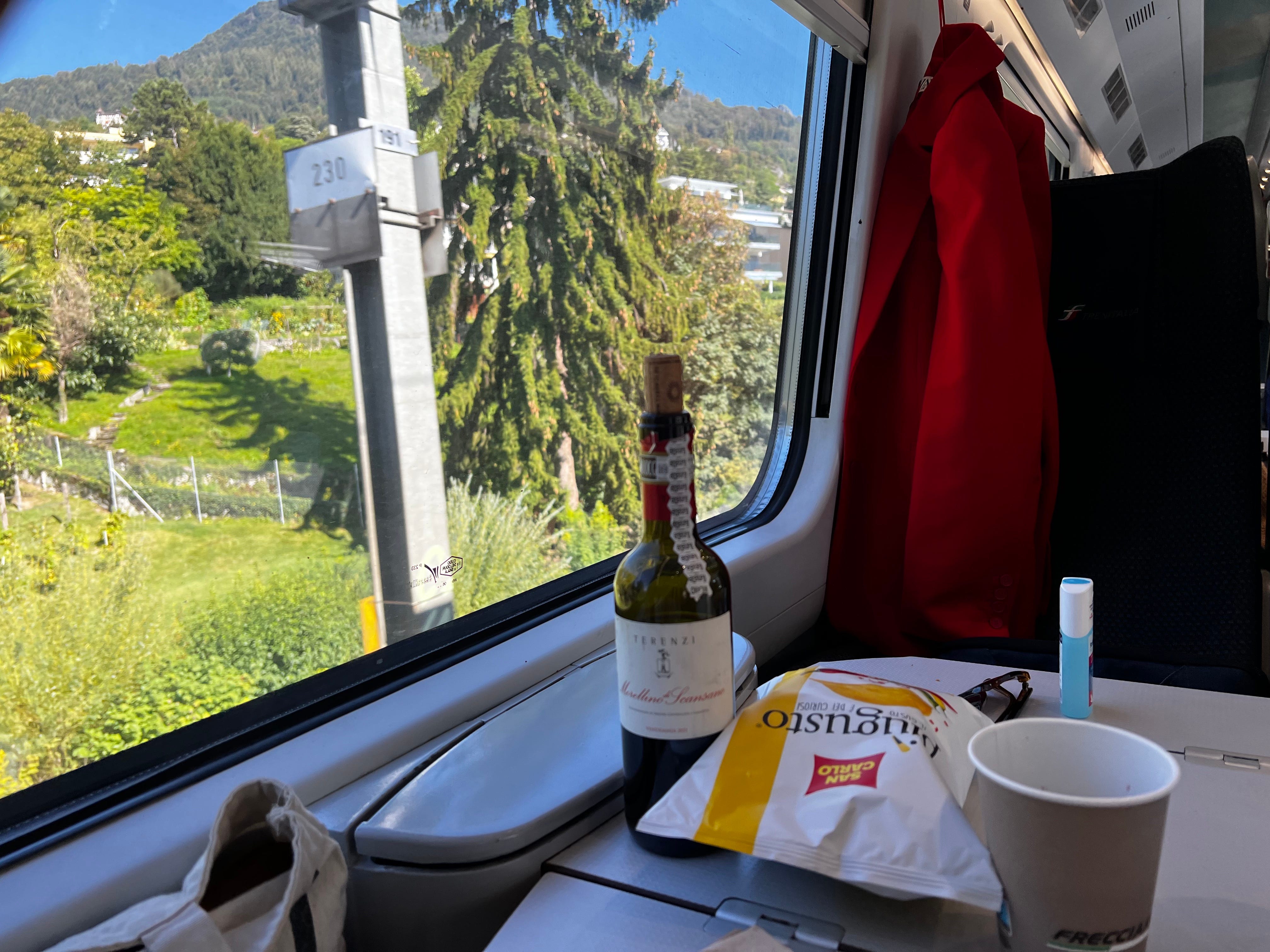 Eine Flasche Wein und eine Tüte Chips auf einem Tisch vor einem Sitzplatz in einem europäischen Zug, der an üppigen Bäumen vorbeifährt