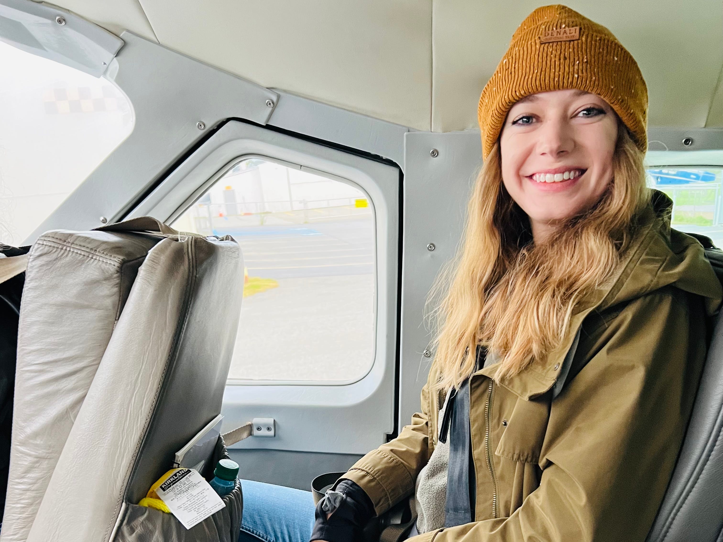 Emily, die einen orangefarbenen Hut, einen beigen Mantel und blaue Jeans trägt, lächelt im Flugzeug.