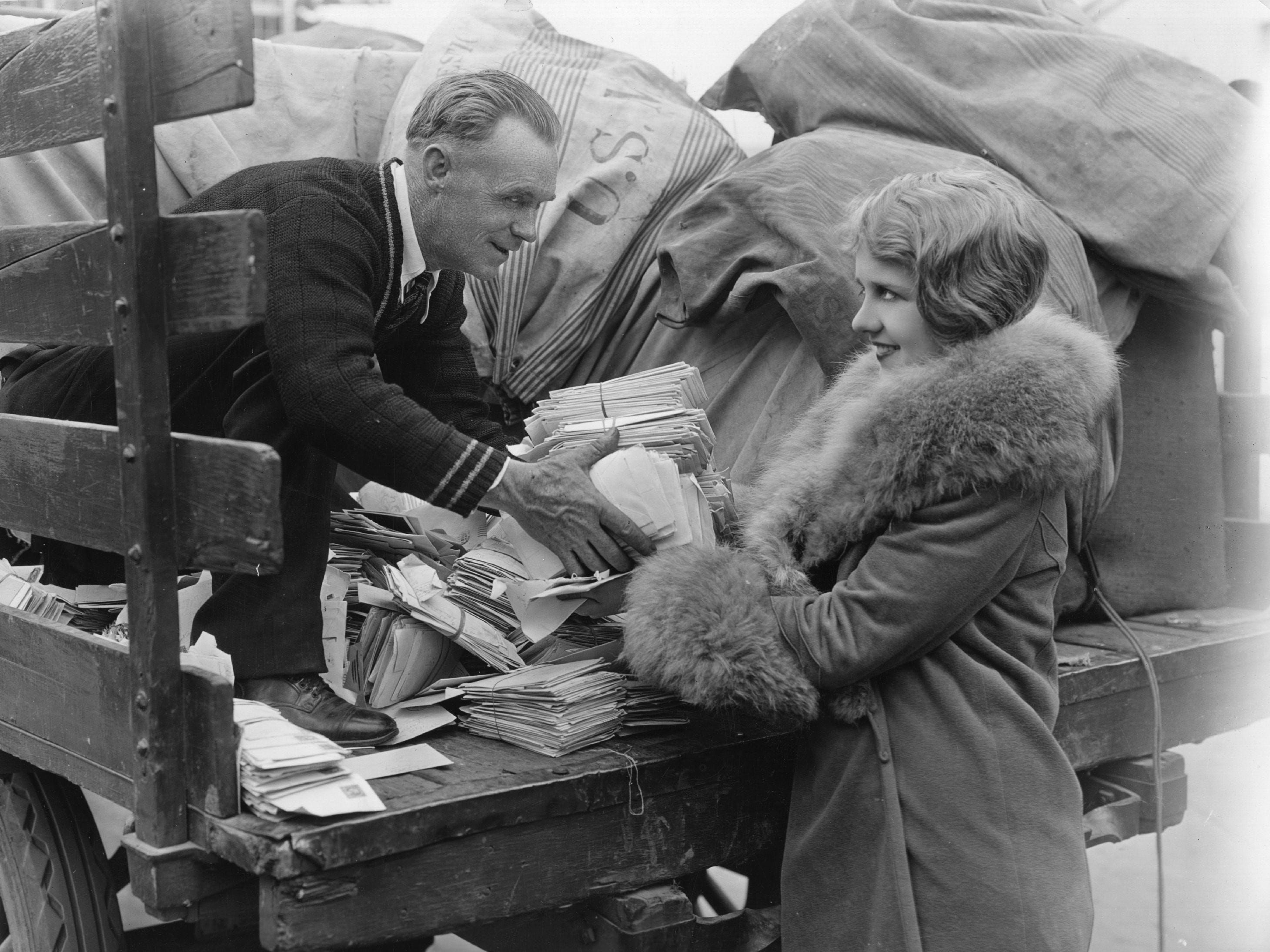 Ein Postangestellter im Anzug überreicht einer Frau im Pelzmantel Post