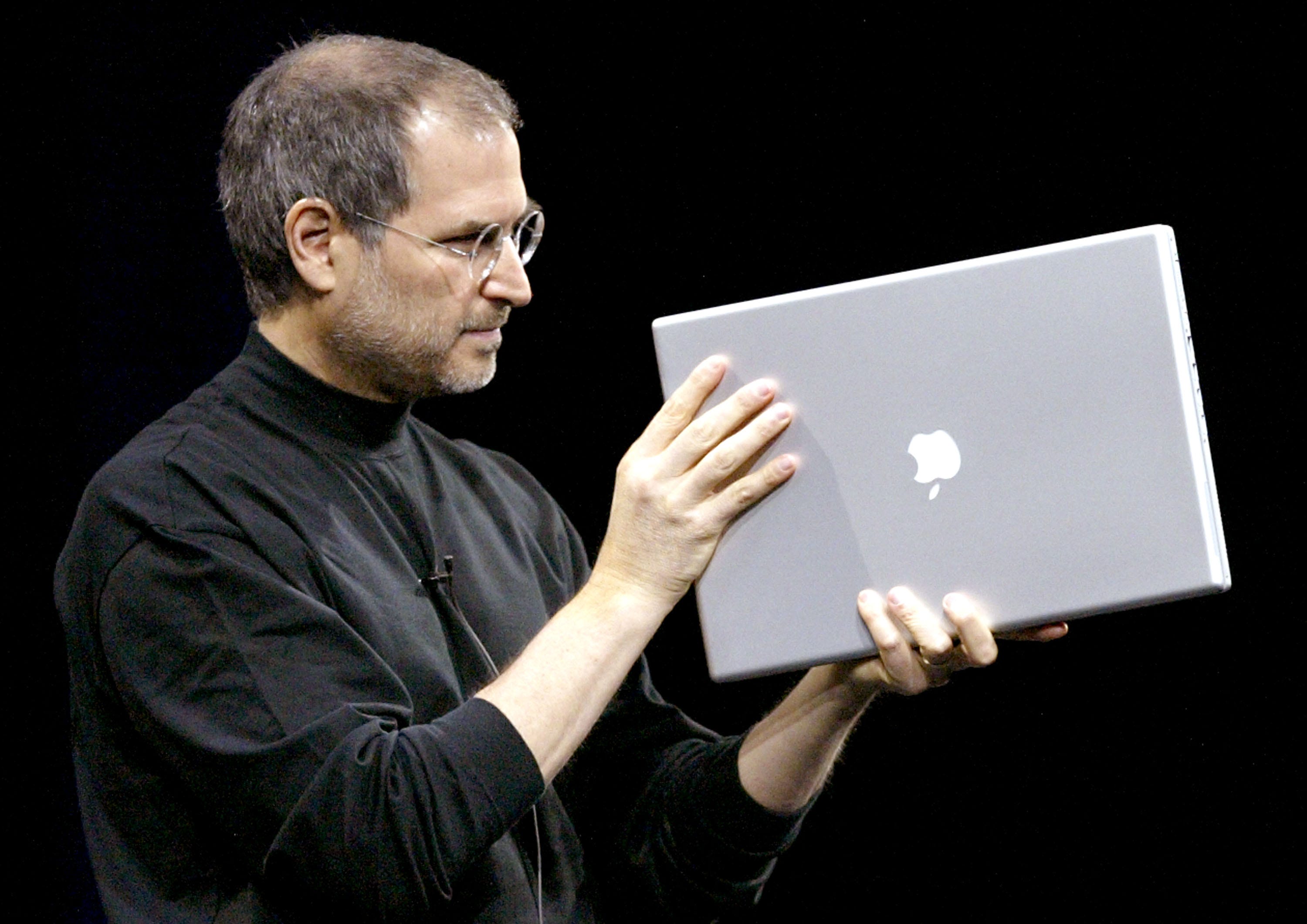 Steve Jobs trägt seinen charakteristischen schwarzen Rollkragenpullover und hält einen Apple-Laptop in der Hand