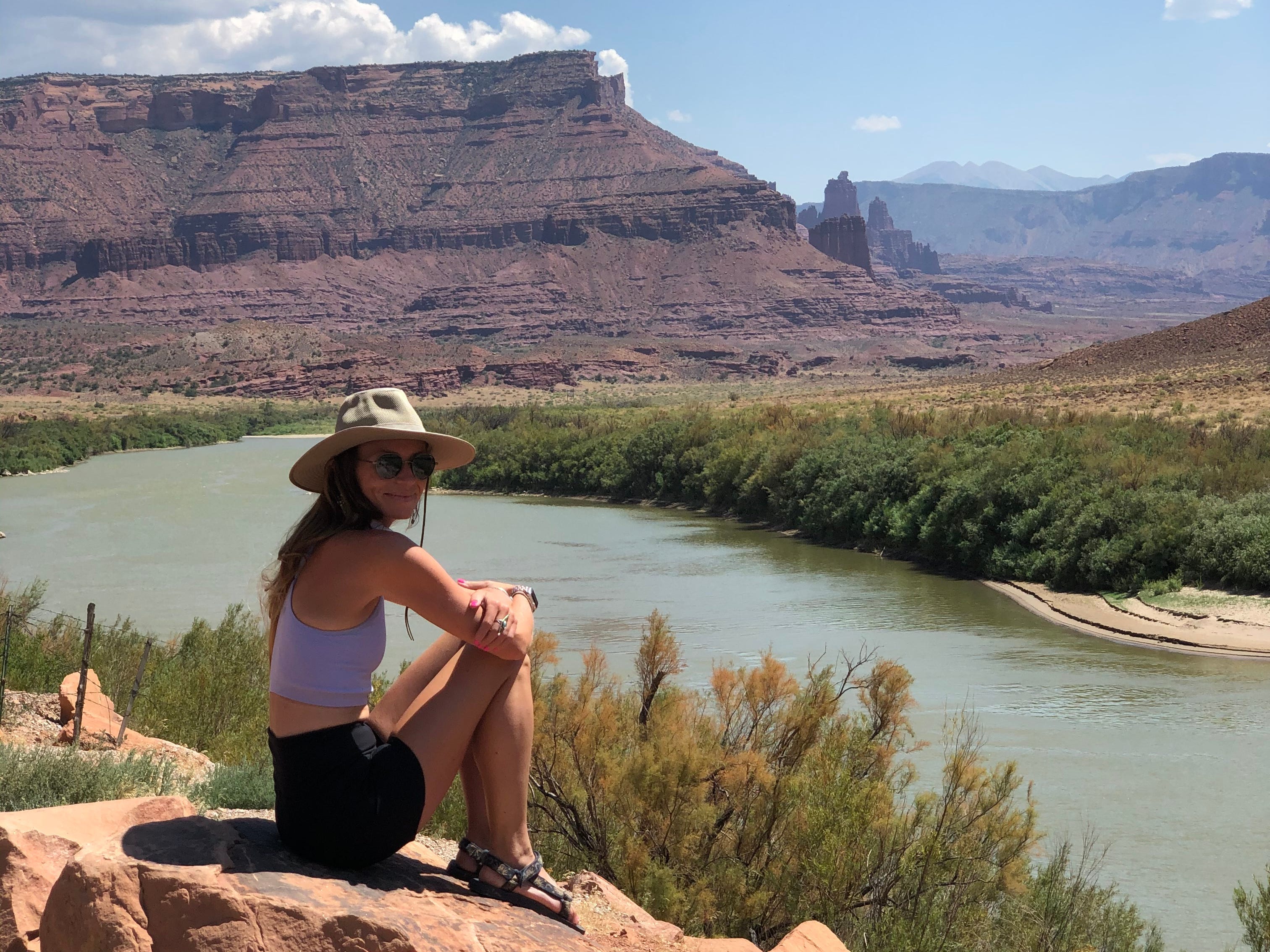 Emily sitzt in einem lila Crop-Top, schwarzen Shorts, einem beigen Hut und einer Sonnenbrille auf einem Felsen und blickt auf ein kleines Gewässer und große Felsformationen.