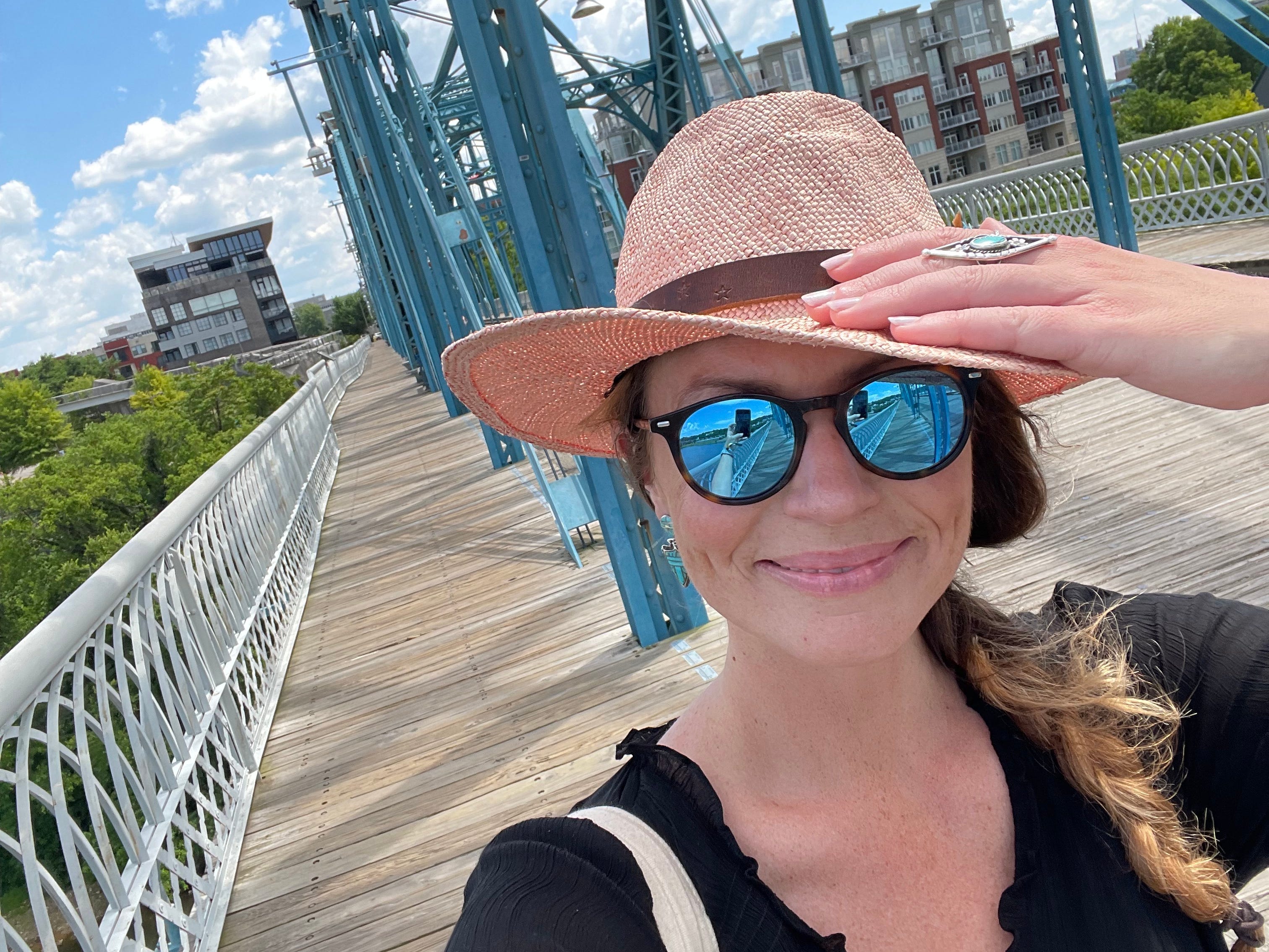 Emily, die ein schwarzes Hemd, eine Sonnenbrille und einen rosa Hut trägt, macht ein Selfie auf einer Brücke.