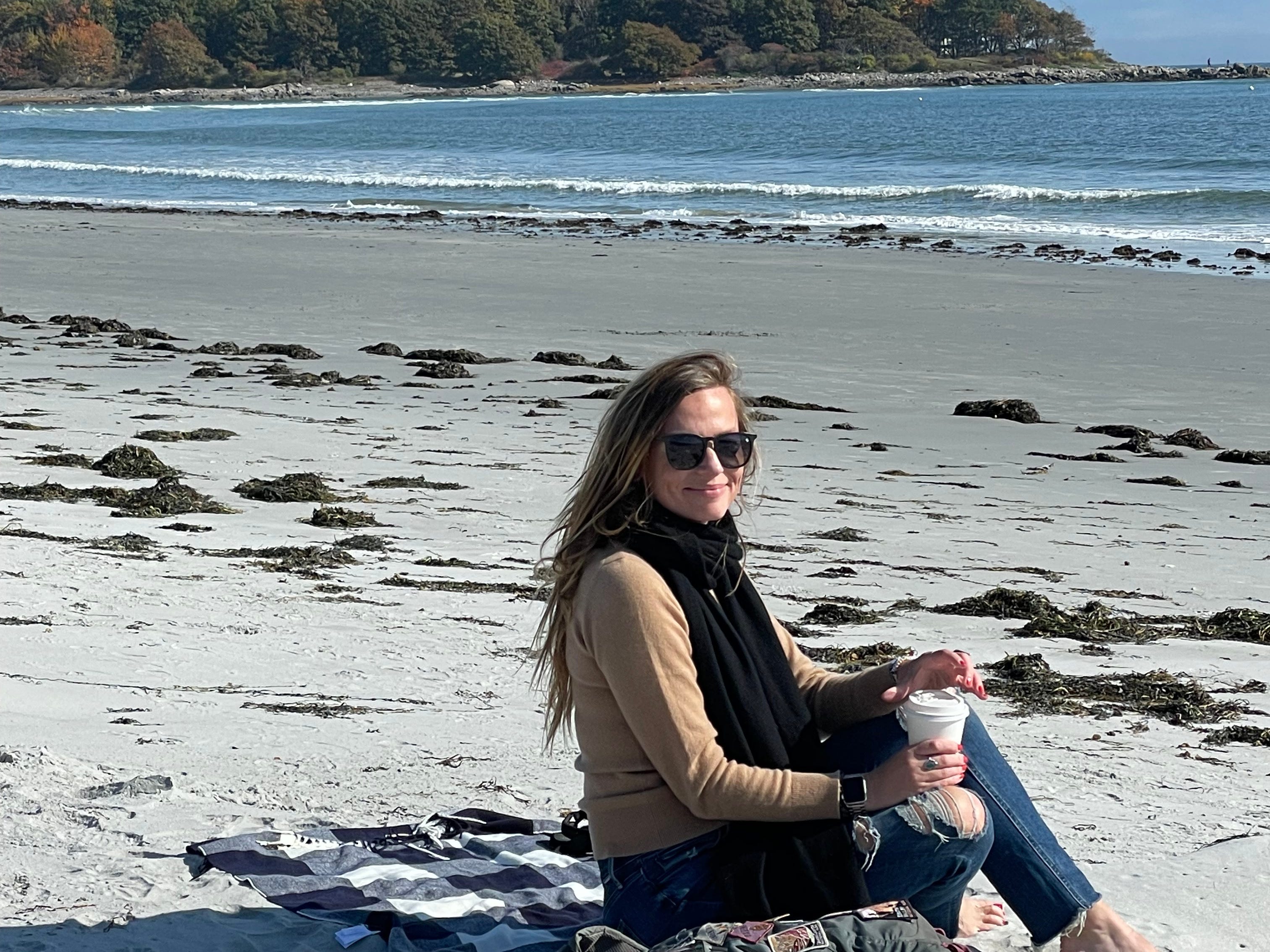 Emily sitzt in einem langärmligen Hemd, einem schwarzen Schal, Jeans und einer Sonnenbrille am Strand und hält eine Kaffeetasse in der Hand.