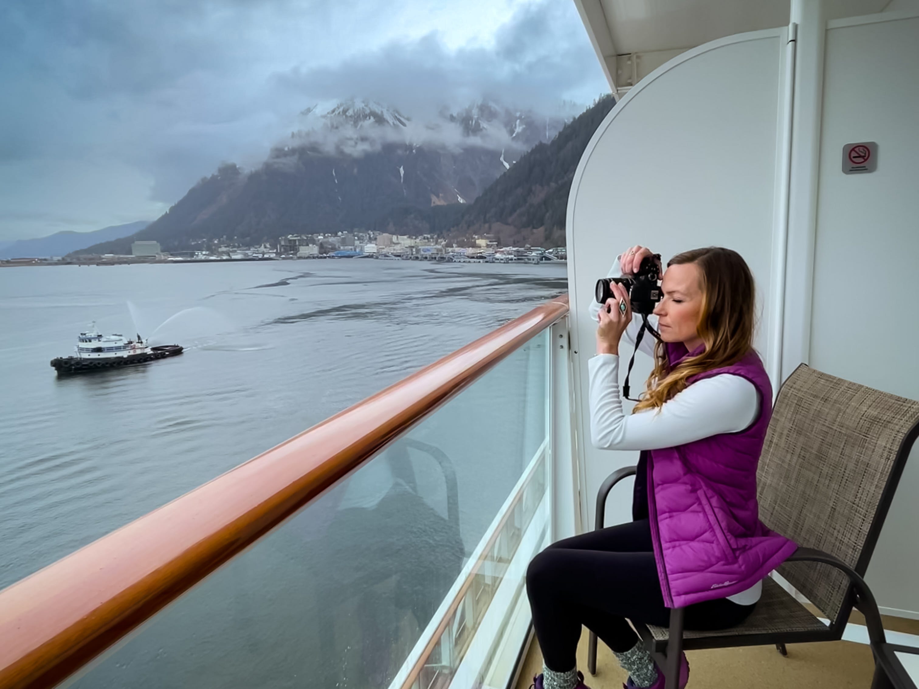 Emily, gekleidet in eine lila Steppweste, ein weißes Langarmhemd, eine schwarze Hose und lila Turnschuhe, macht ein Foto auf dem Balkon einer Alaska-Kreuzfahrt.