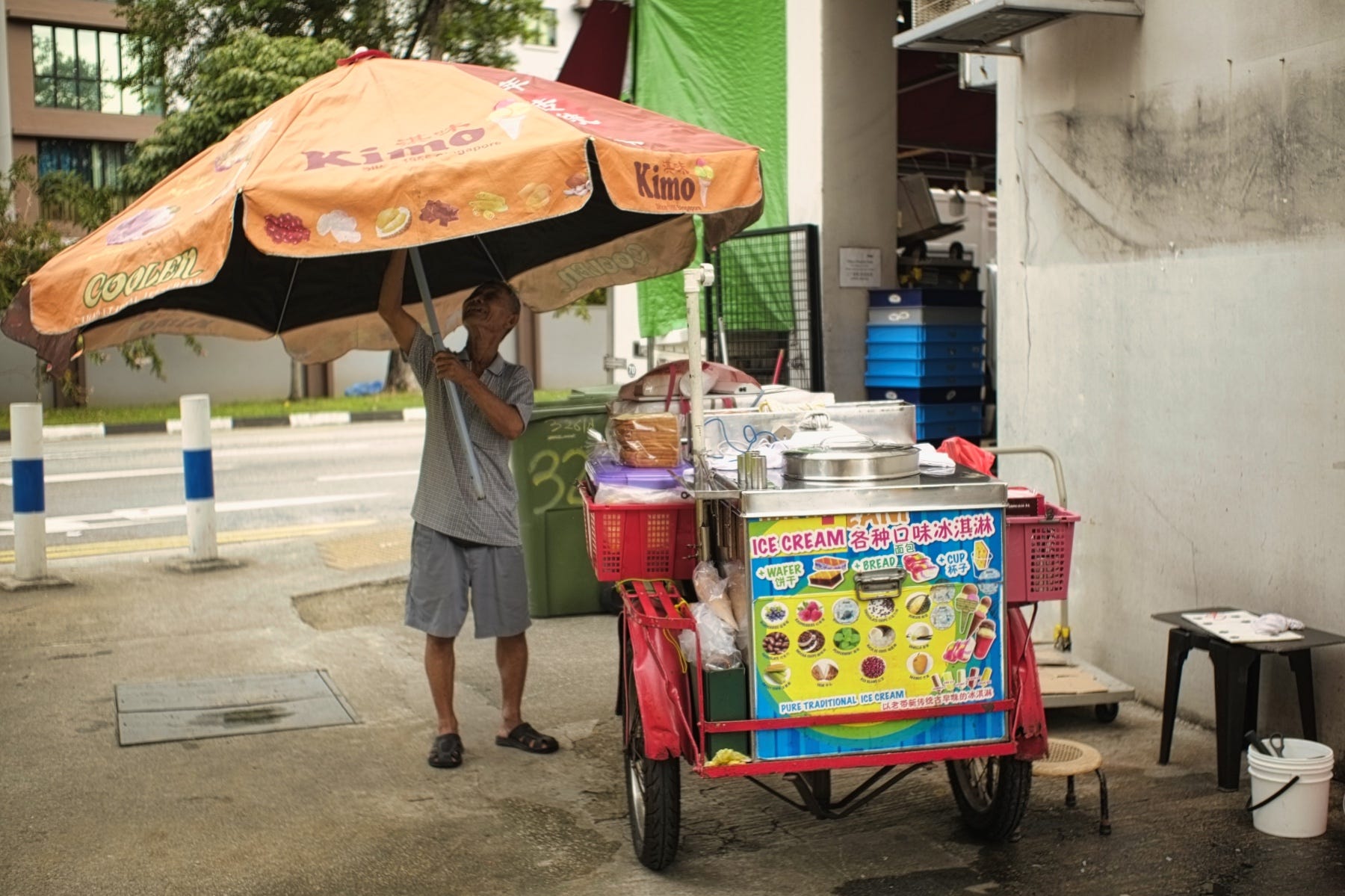 Ein älterer singapurischer Mann mit schlanker Statur hält einen riesigen Regenschirm neben seinem Eiswagen.