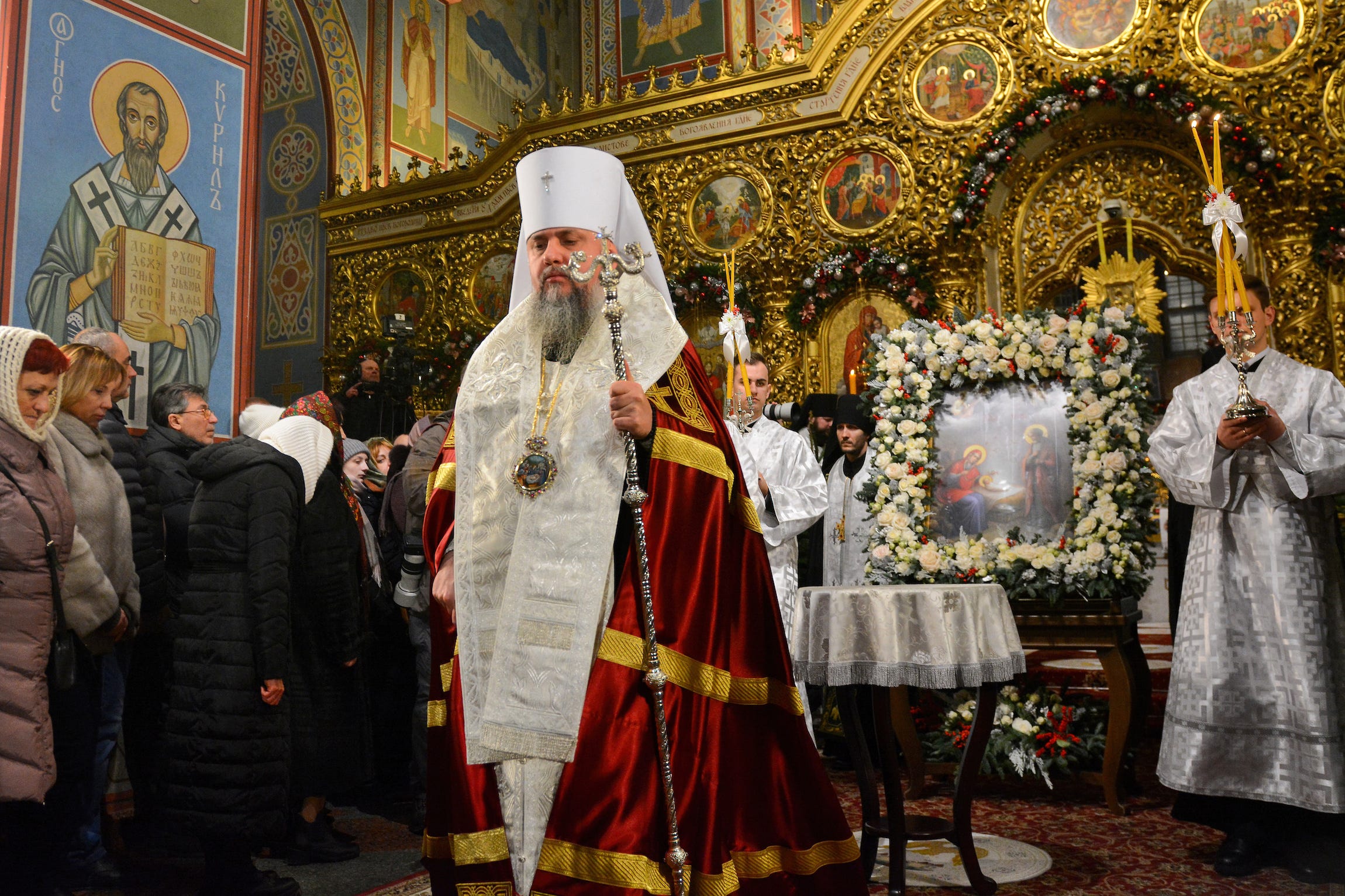 Ein orthodoxer Priester in zeremoniellen Gewändern aus Weiß, Rot und Gold hält ein Zepter und leitet einen Gottesdienst in einer goldgeschmückten Kirche in Kiew, Ukraine.