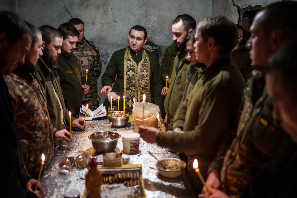 Kaplan Ivan von der 72. Brigade der ukrainischen Streitkräfte hält am 24. Dezember 2023 einen Gottesdienst im Oblast Donezk, Ukraine.  Man sieht ihn am Kopfende eines Tisches stehen, umgeben von Soldaten in einem Betonraum, von denen einige Stabkerzen um Grundnahrungsmittel halten.
