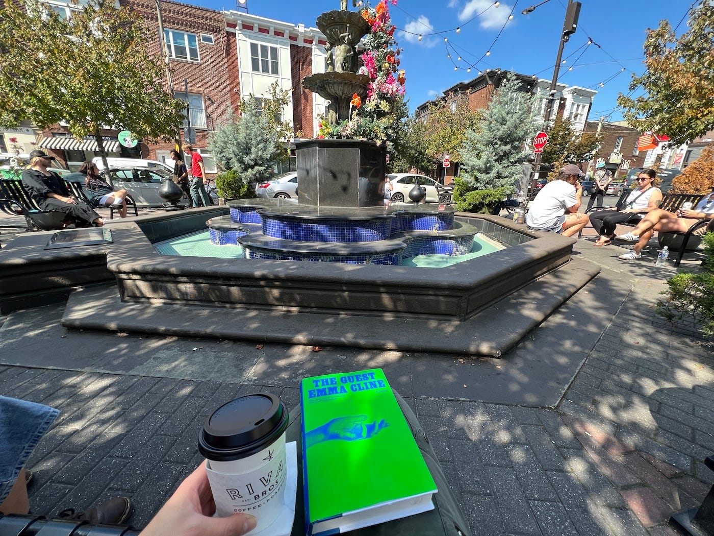 Eine Frau hält eine Tasse Kaffee zum Mitnehmen und ein Buch auf ihrem Schoß auf einer Bank vor einem Brunnen im Sonnenschein.