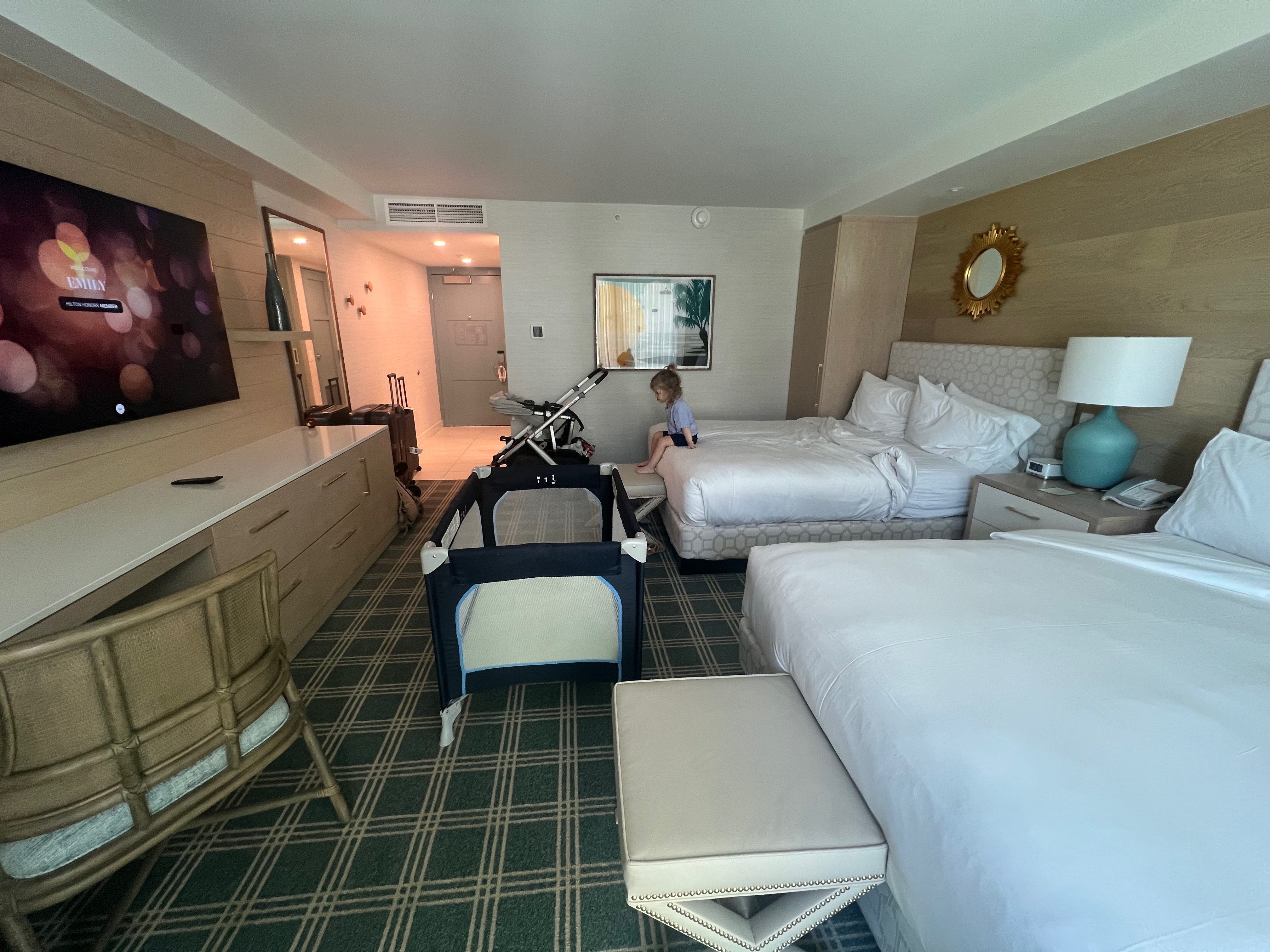 Ein Hotelzimmer mit zwei Betten, auf dem ein Kind sitzt, einem Reisebett, einem Schreibtisch und Gepäck.