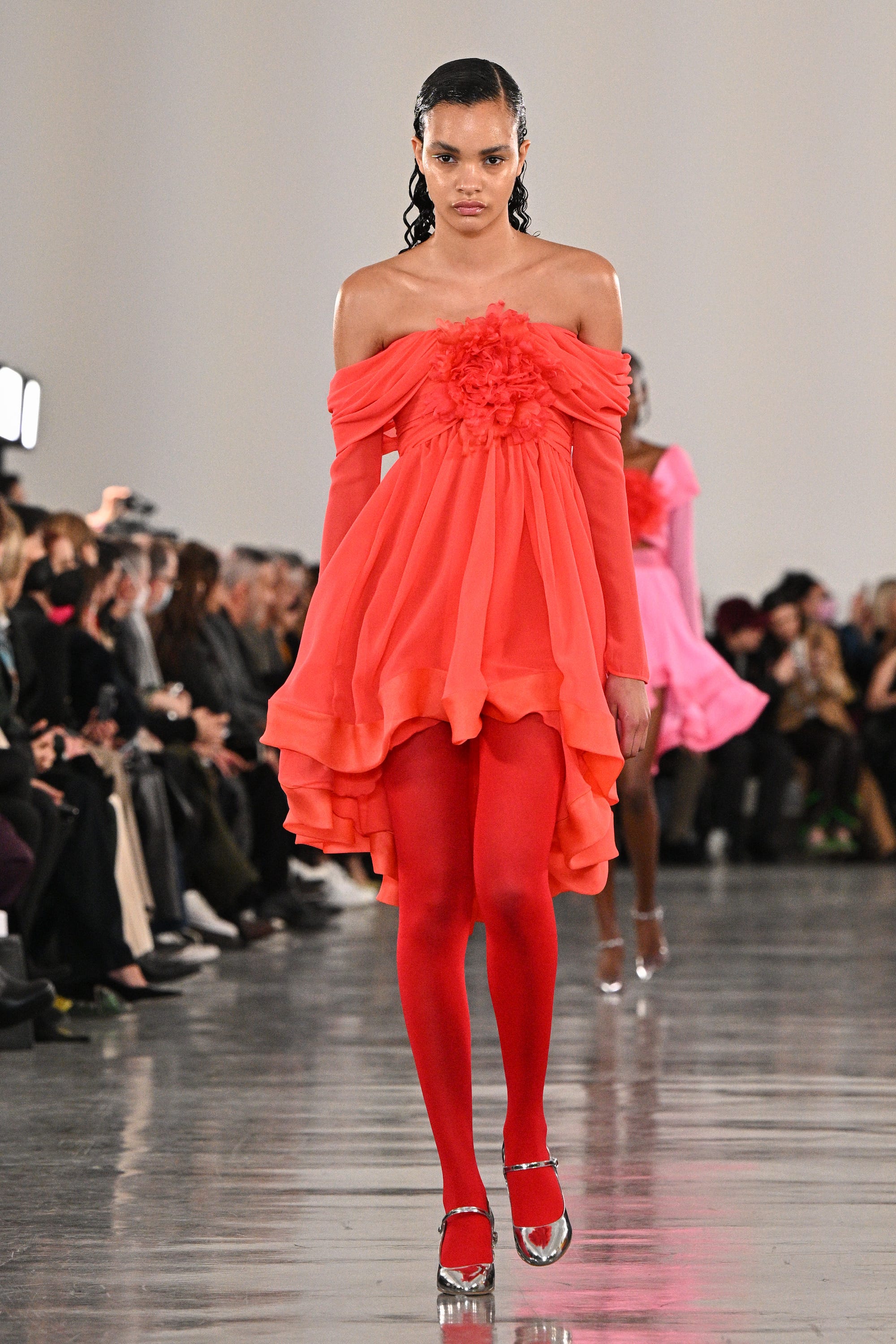 Ein Model läuft über einen Laufsteg und trägt ein schulterfreies rotes Kleid mit roten Strumpfhosen und silbernen Schuhen.