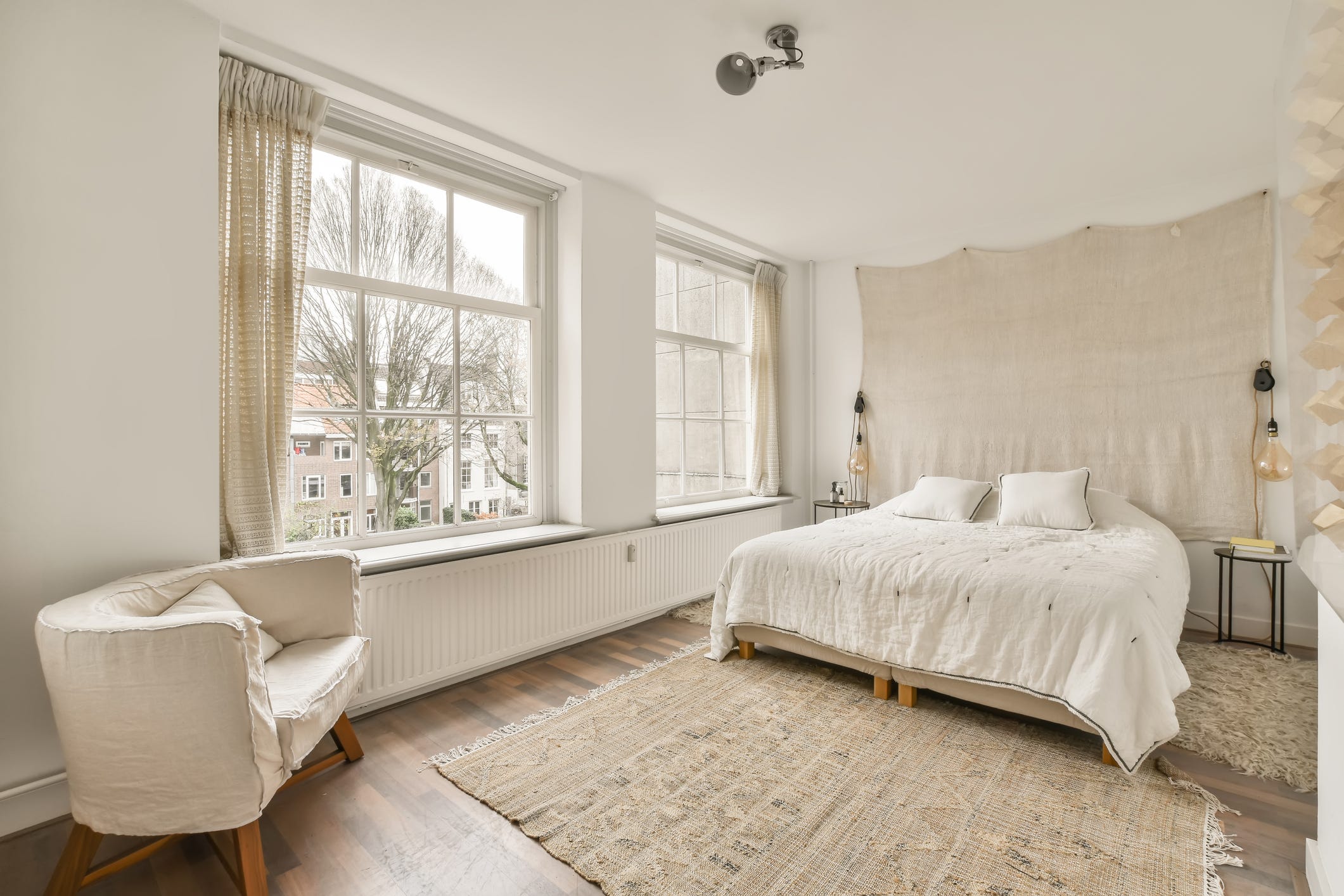 Beiges Zimmer mit großen Glasfenstern, weißem Bett, beigem Wandteppich, beigem Teppich und beigem Stuhl