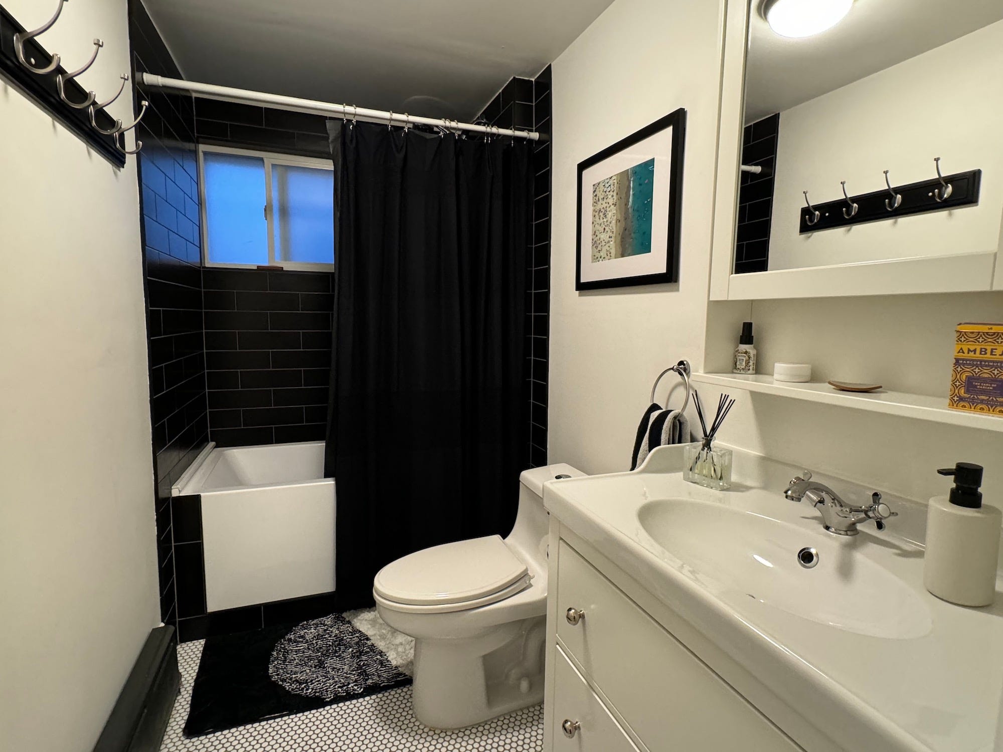 Ein Badezimmer mit einer schwarz gefliesten Badewanne und einer weißen Toilette, einem Waschbecken und einem Waschtisch mit Diffusor und Seifenspender darauf.