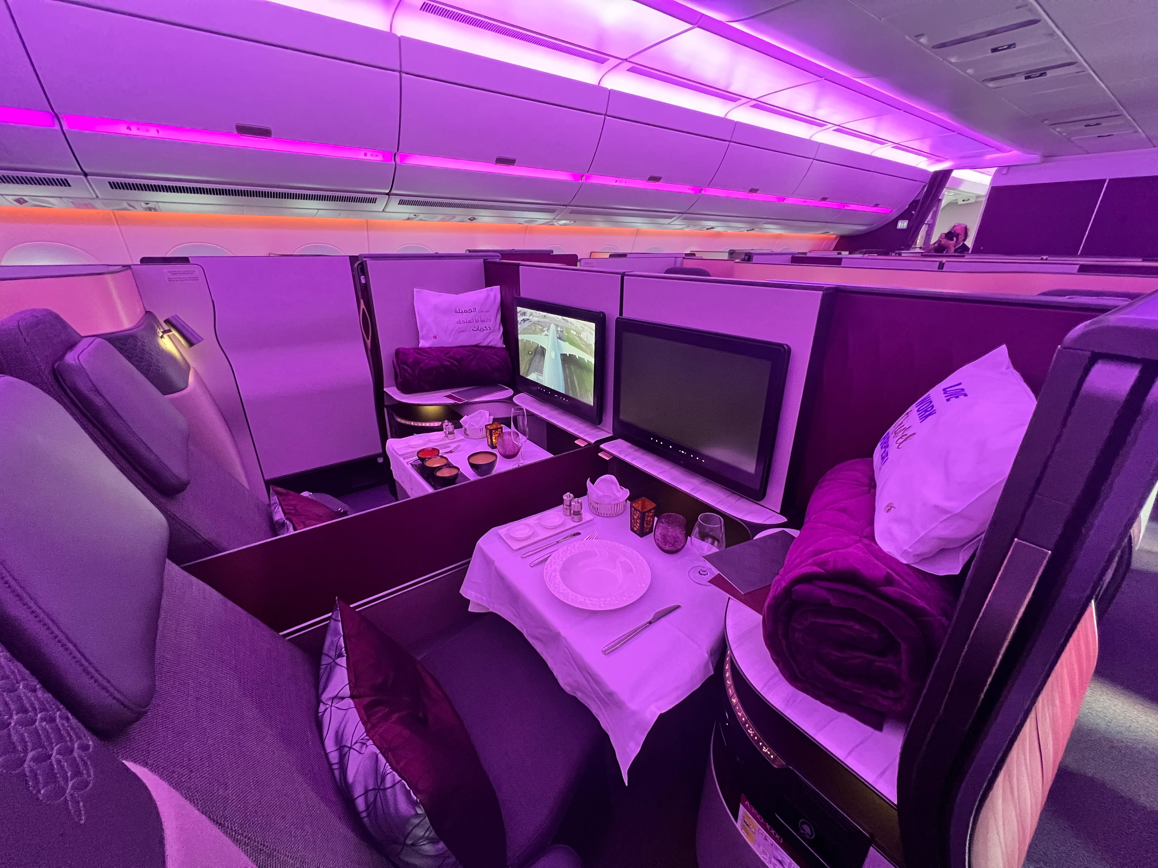 Ein Blick auf zwei nebeneinander liegende Business-Class-Sitze von Qatar Airways mit violettem Farbton und bereitgelegtem Geschirr.