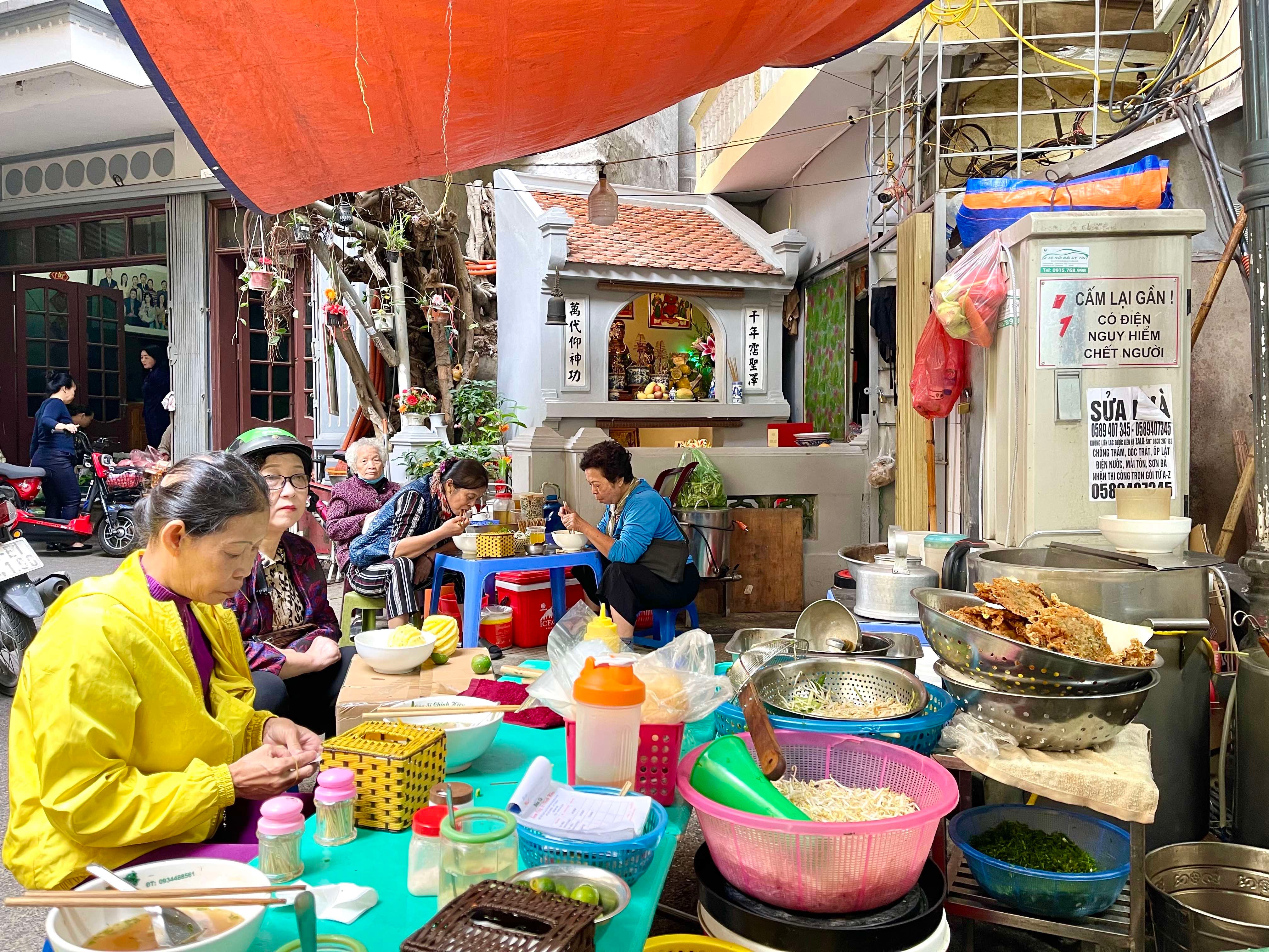 Frauen essen auf Hockern vor der Straßenküche Bún Cá Sâm Cây Si neben dem Schrein in Hanoi