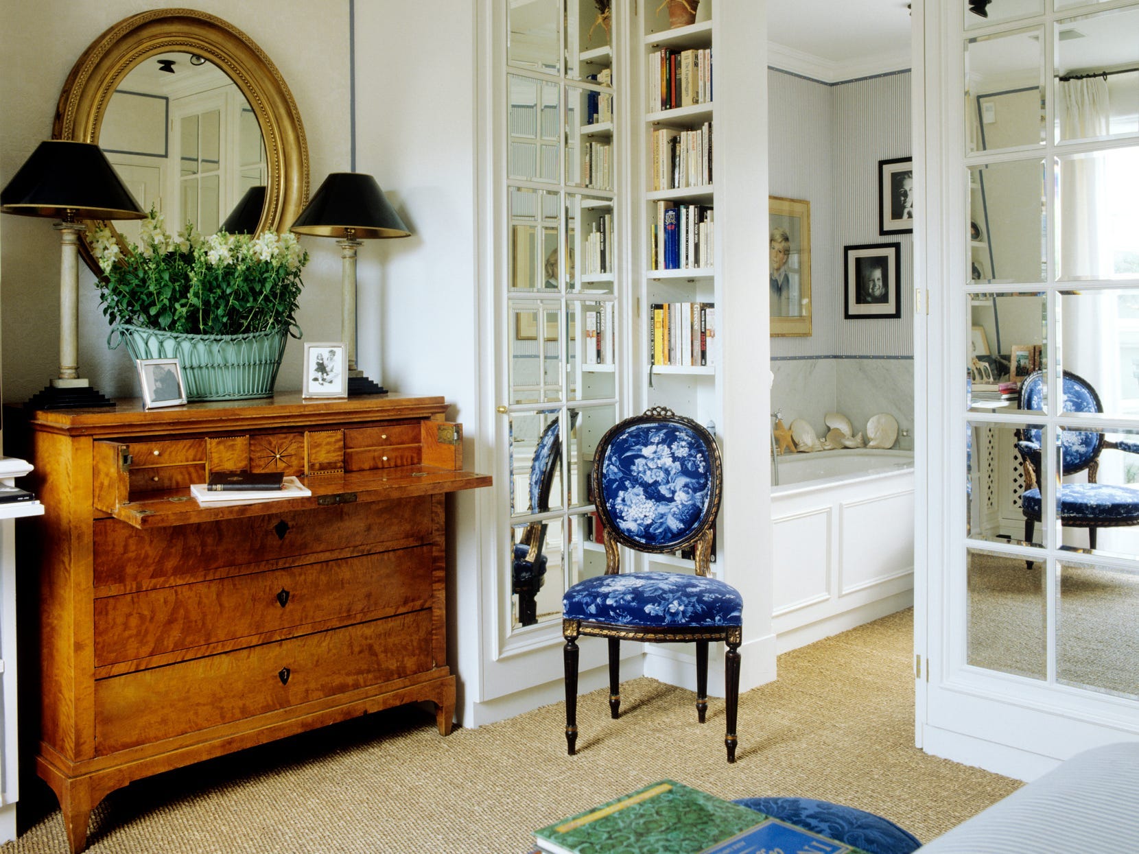 Eine Vintage-Kommode aus Holz mit einem ausklappbaren Regal, darunter ein Spiegel, Blumen und zwei Lampen.  Neben der Kommode stehen ein blauer Stuhl, ein kleines Bücherregal und ein Eingang zu einem Badezimmer.