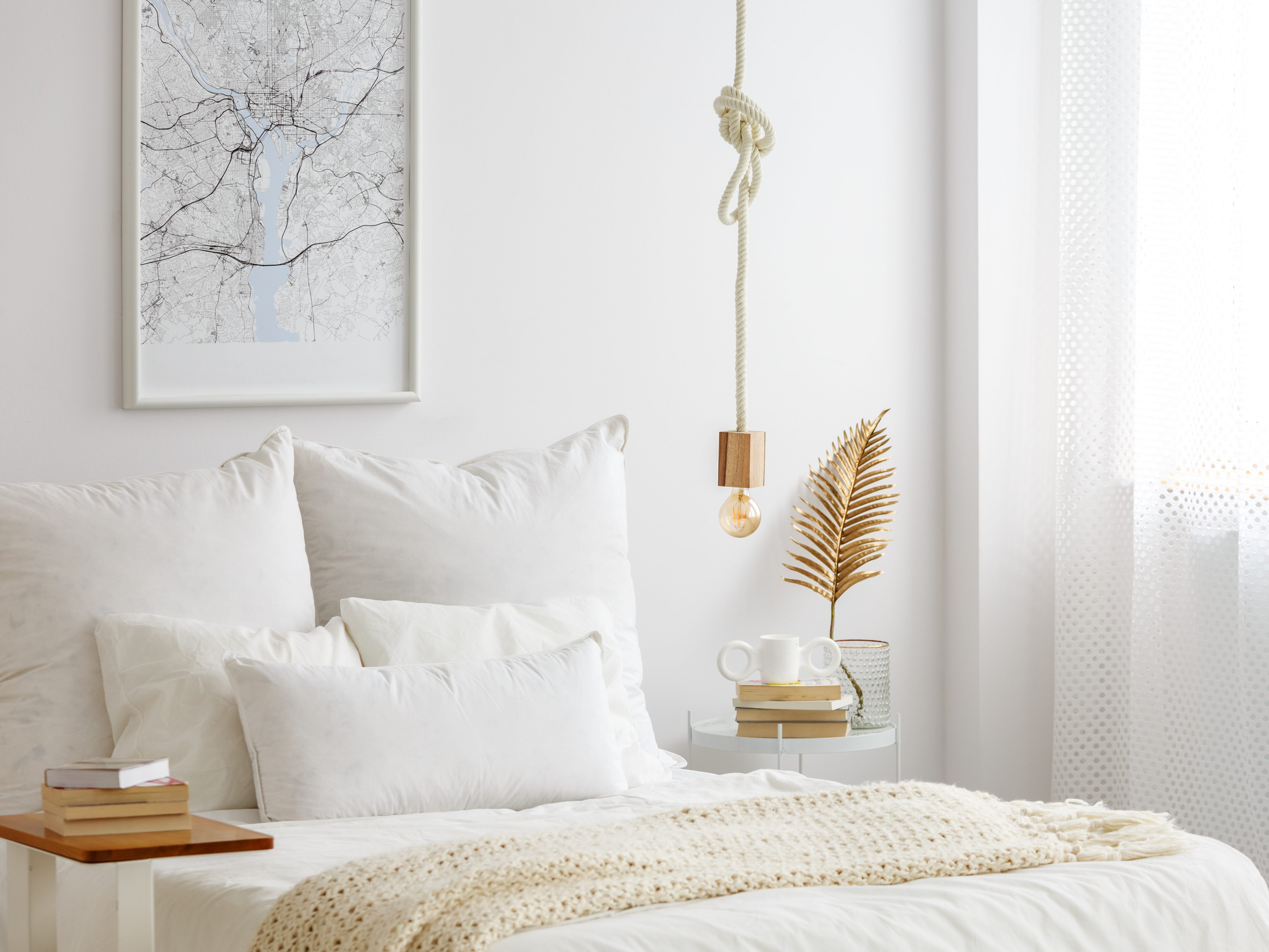 Ein Schlafzimmer mit weißen Wänden, weißen Vorhängen und weißer Bettwäsche.  Über dem Bett hängt eine Glühbirne an einem Seil und neben dem Bett steht ein Beistelltisch mit Goldakzenten.
