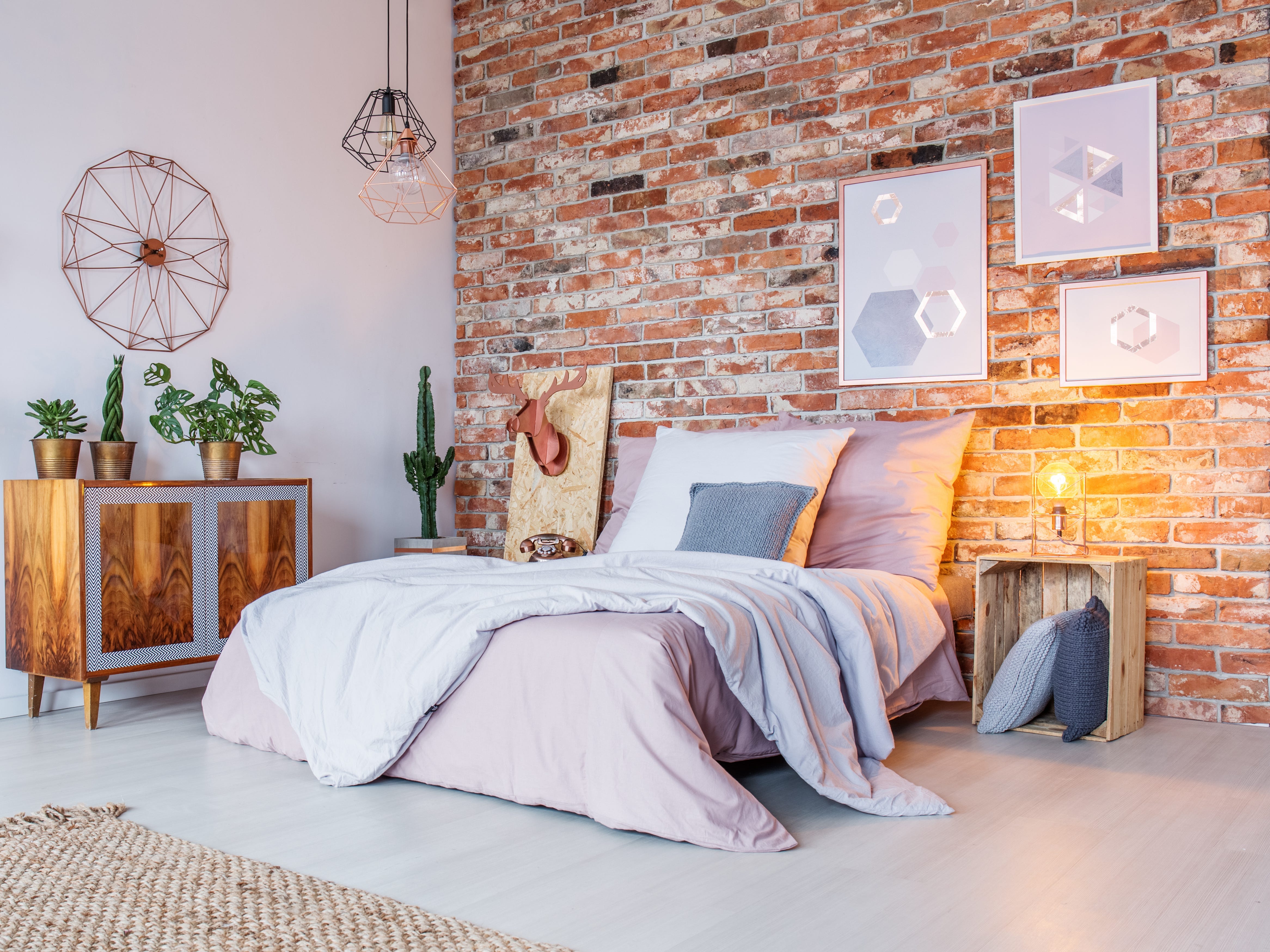Ein helles Schlafzimmer mit einer unverputzten Ziegelwand, einem Bett mit einer hellrosa Bettdecke, einer Holzkommode und vielen dekorativen Wandelementen.