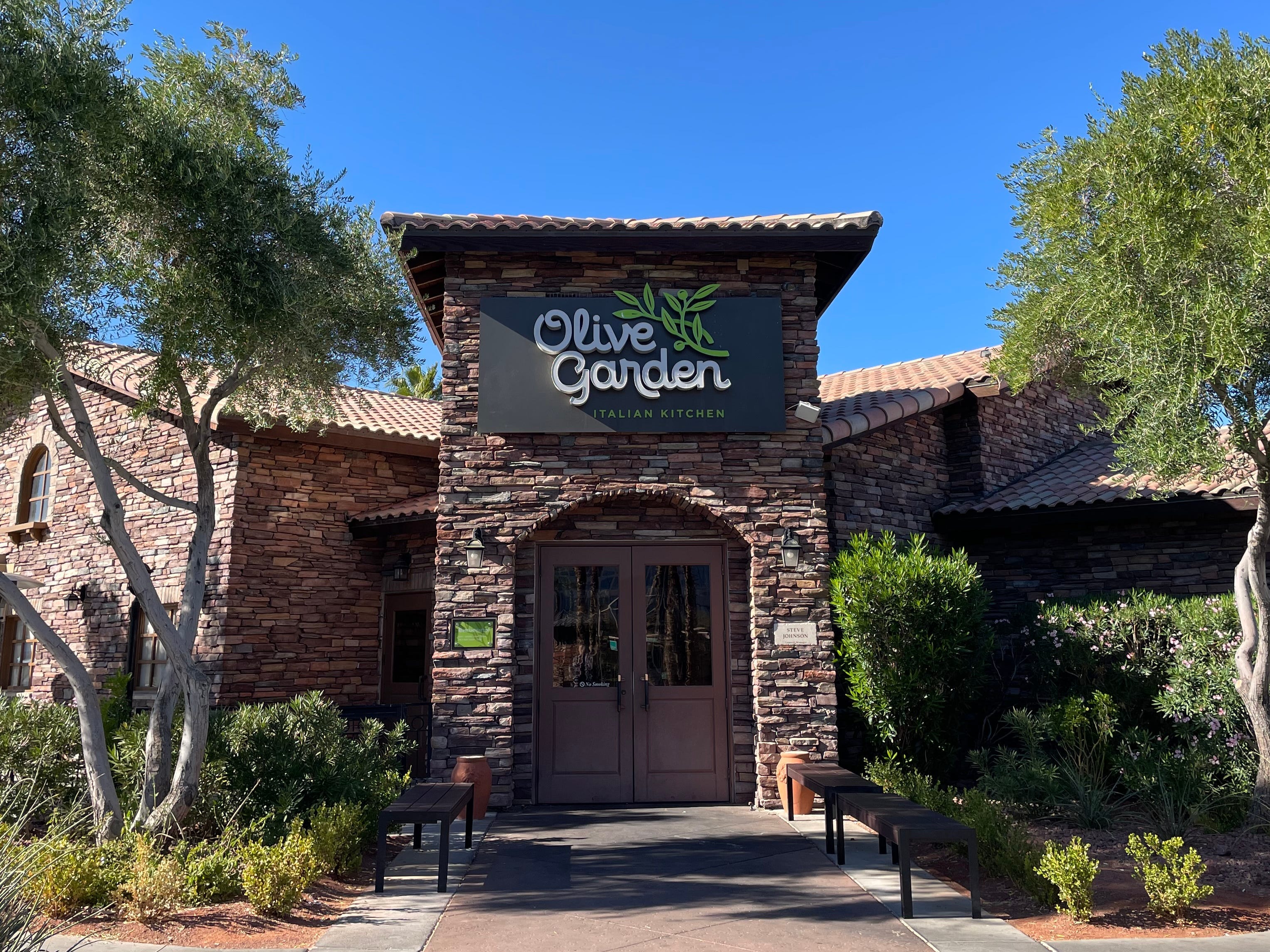 Außenansicht des Olivengartens.  Das Backsteingebäude mit dem „Olive Garden“-Schild ist von Bäumen und einem strahlend blauen Himmel umgeben