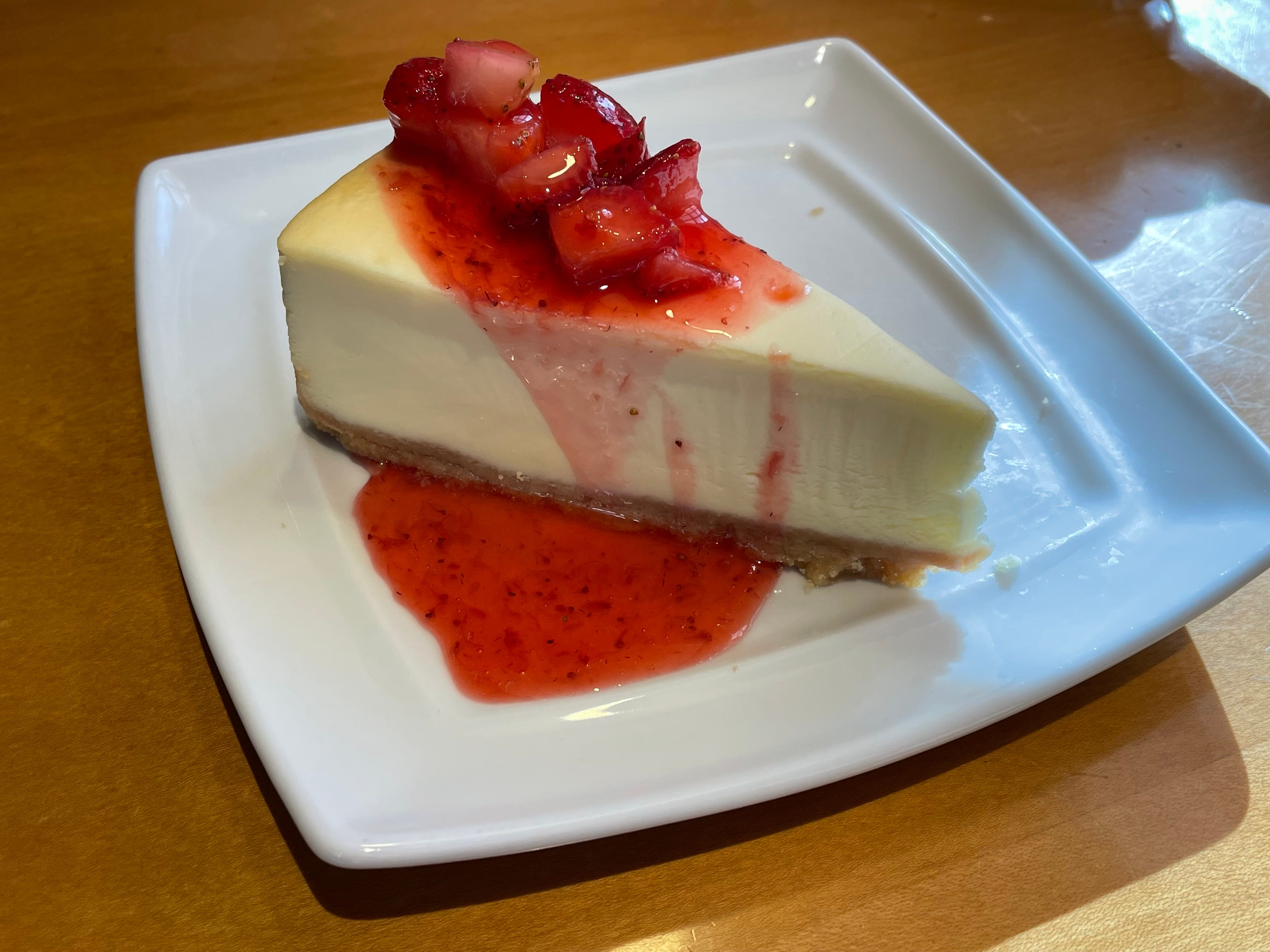 Ein Stück Käsekuchen mit Erdbeersauce und einem Stapel Erdbeeren darauf liegt auf einem quadratischen weißen Teller auf einem braunen Tisch im Olive Garden