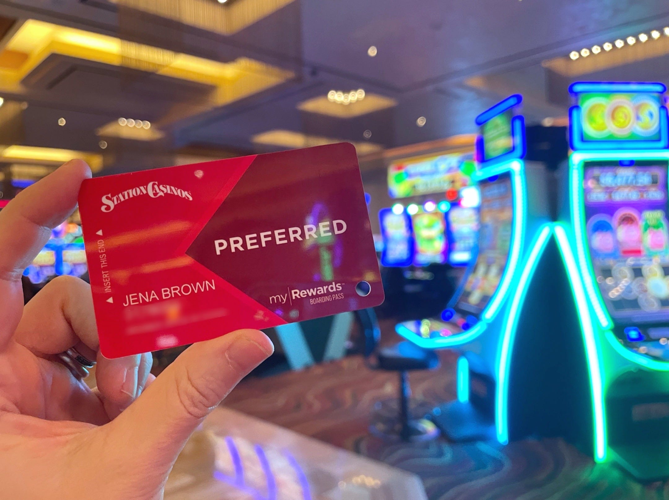 Eine Hand hält eine Prämienkarte von Station Casinos hoch, während sie in einem hell erleuchteten Raum vor einer Reihe von Casino-Automaten steht.