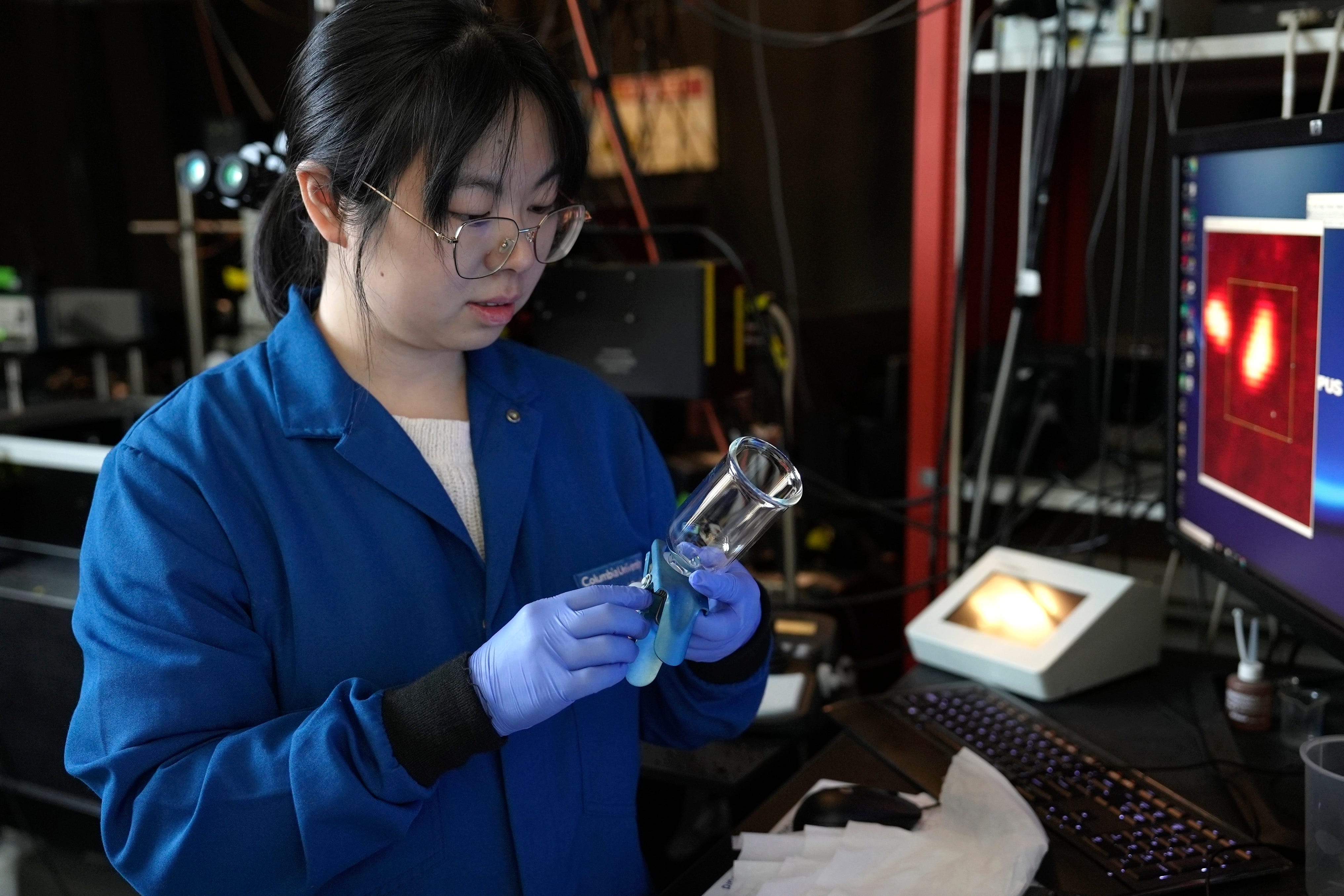 Eine Wissenschaftlerin trägt blaue Handschuhe, während sie eine Probe in einem durchsichtigen Glas betrachtet, die sie gerade unter ein Mikroskop legt.