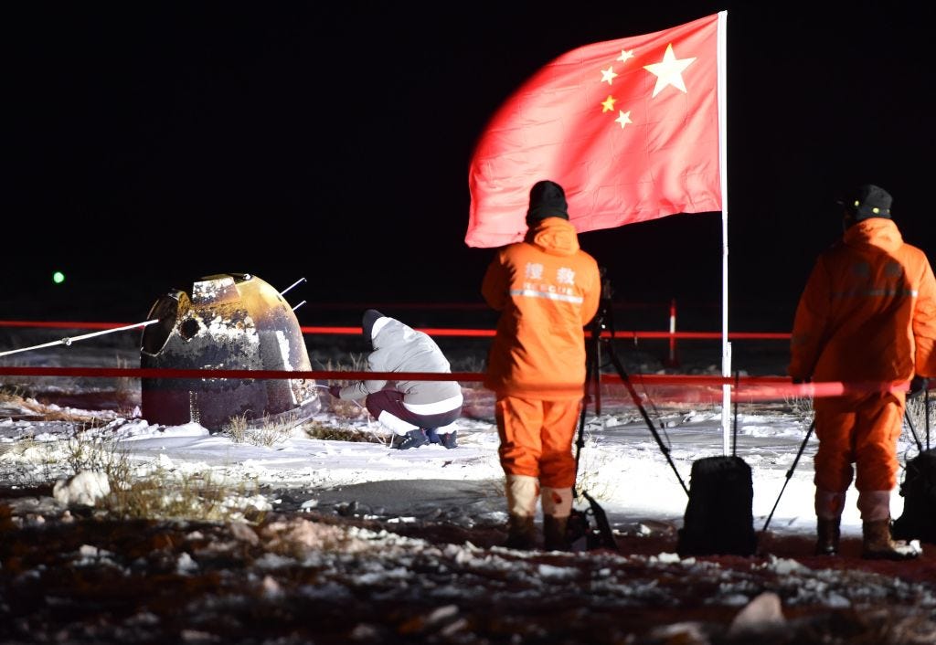 Ein Bild zeigt die verbrannte Kapsel der chinesischen Mission Chang'e 5, die nachts auf einem verschneiten Feld liegt.  Im Vordergrund stehen eine chinesische Flagge und zwei wärmend gekleidete Arbeiter in orangefarbener Arbeitskleidung.  Jemand inspiziert die Sonde, die sich in einem abgesperrten Bereich befindet.