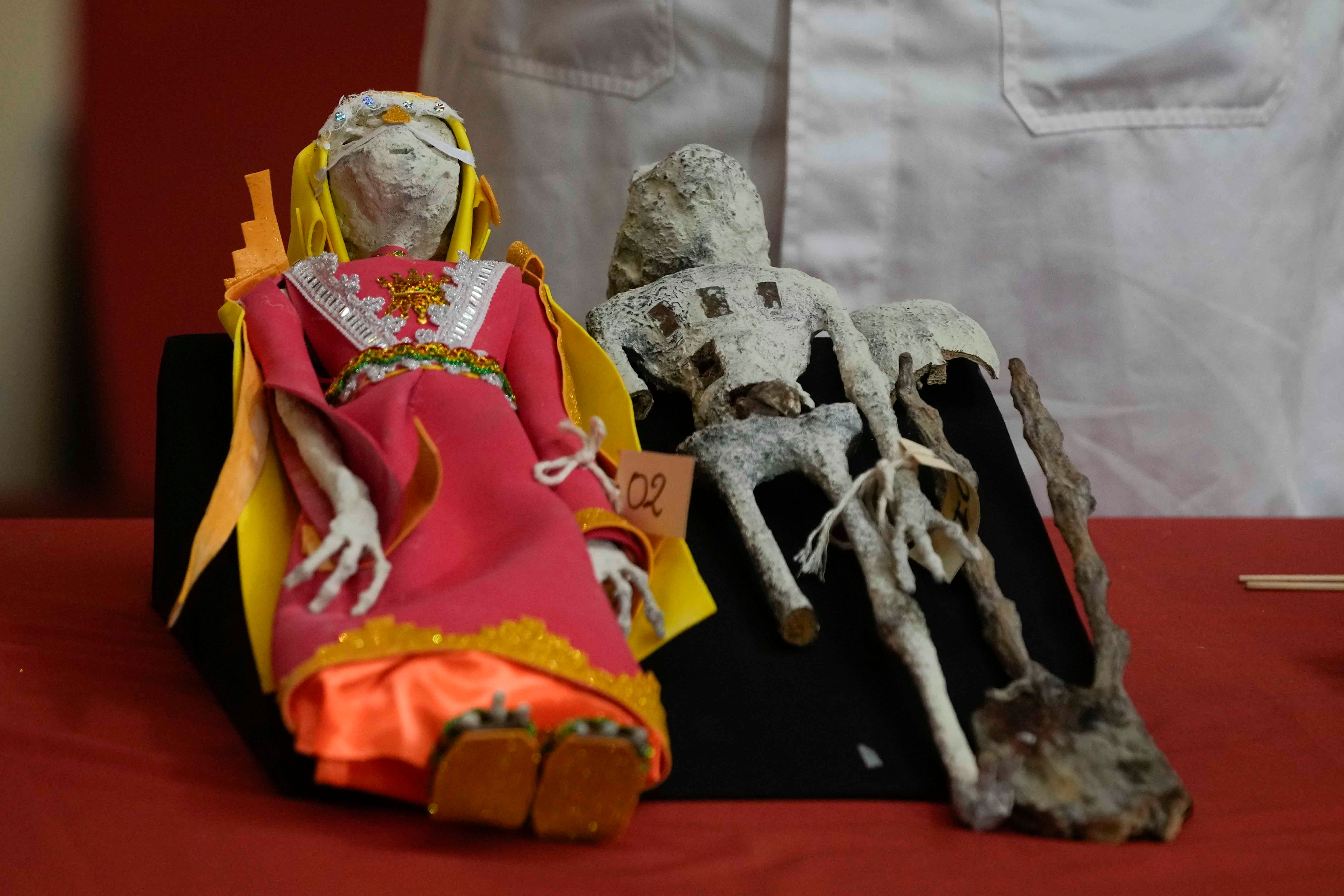 Auf einem Tisch mit roter Tischdecke sind zwei humanoide Puppen aus Papier, Leim sowie menschlichen und tierischen Knochen ausgestellt.