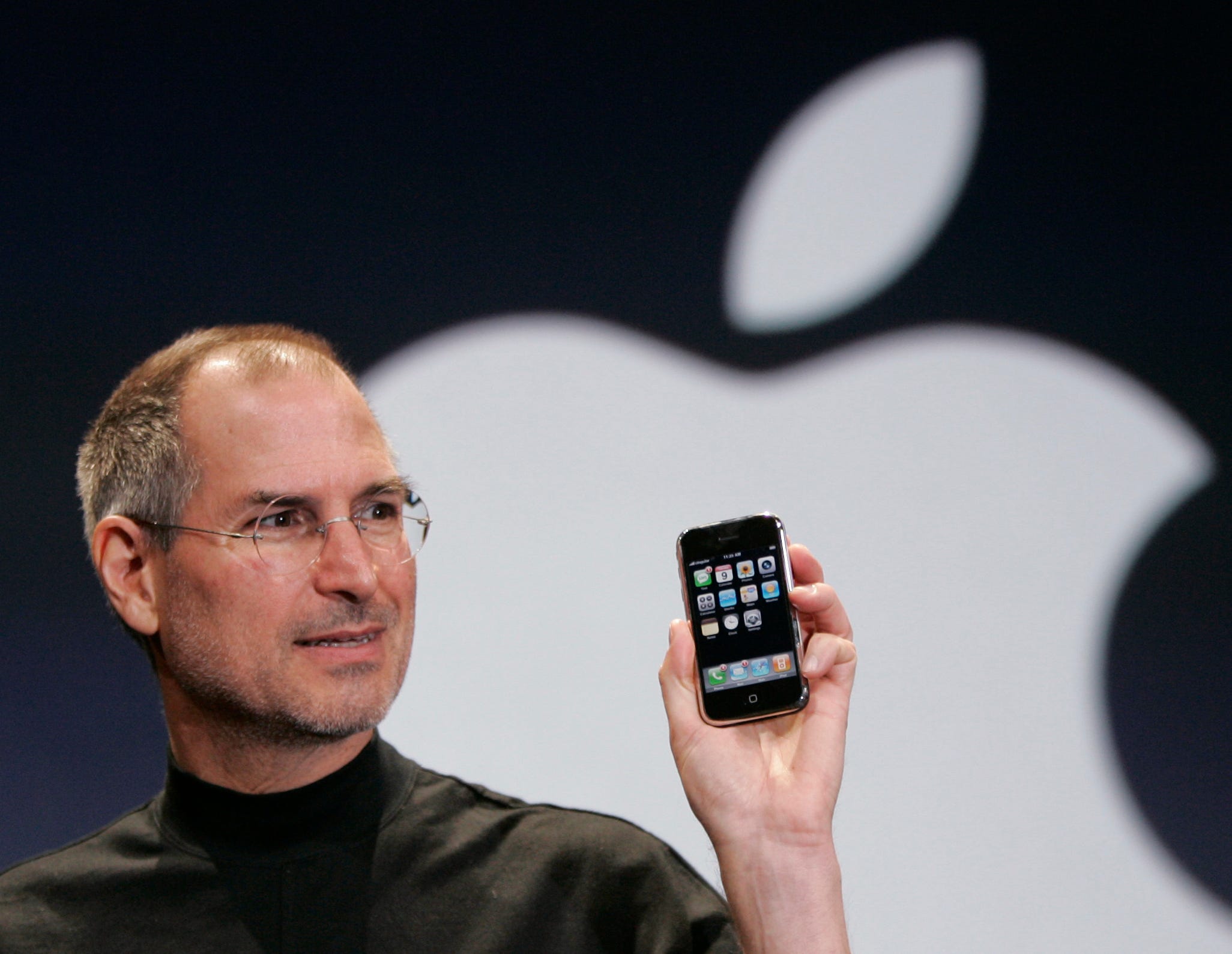 Der frühere Apple-Chef Steve Jobs präsentiert auf der Bühne einer Apple-Veranstaltung das erste iPhone, im Hintergrund taucht das Apple-Logo auf.