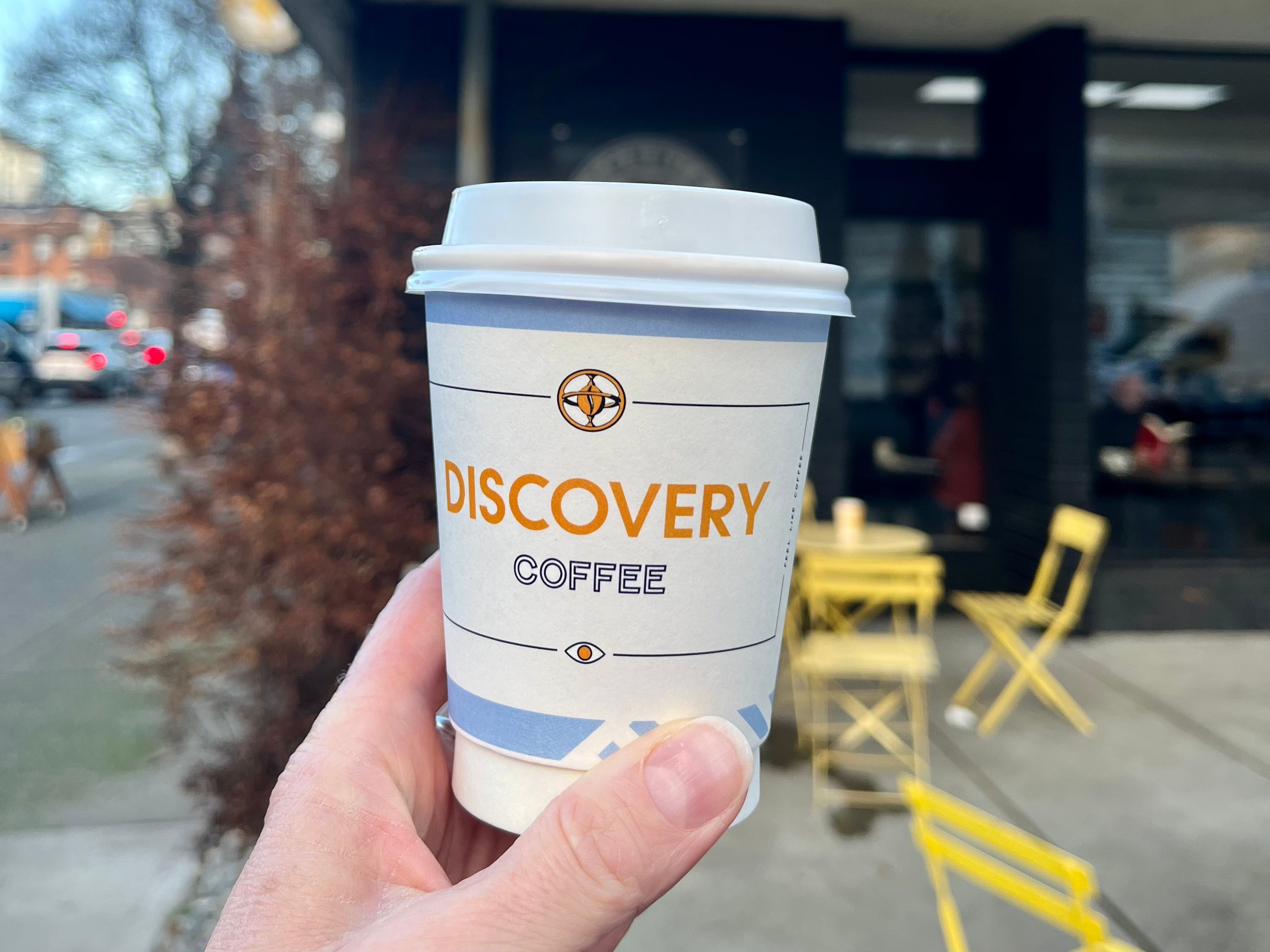 Hand hält eine Tasse Entdeckungskaffee zum Mitnehmen vor dem Entdeckungskaffeecafé hoch
