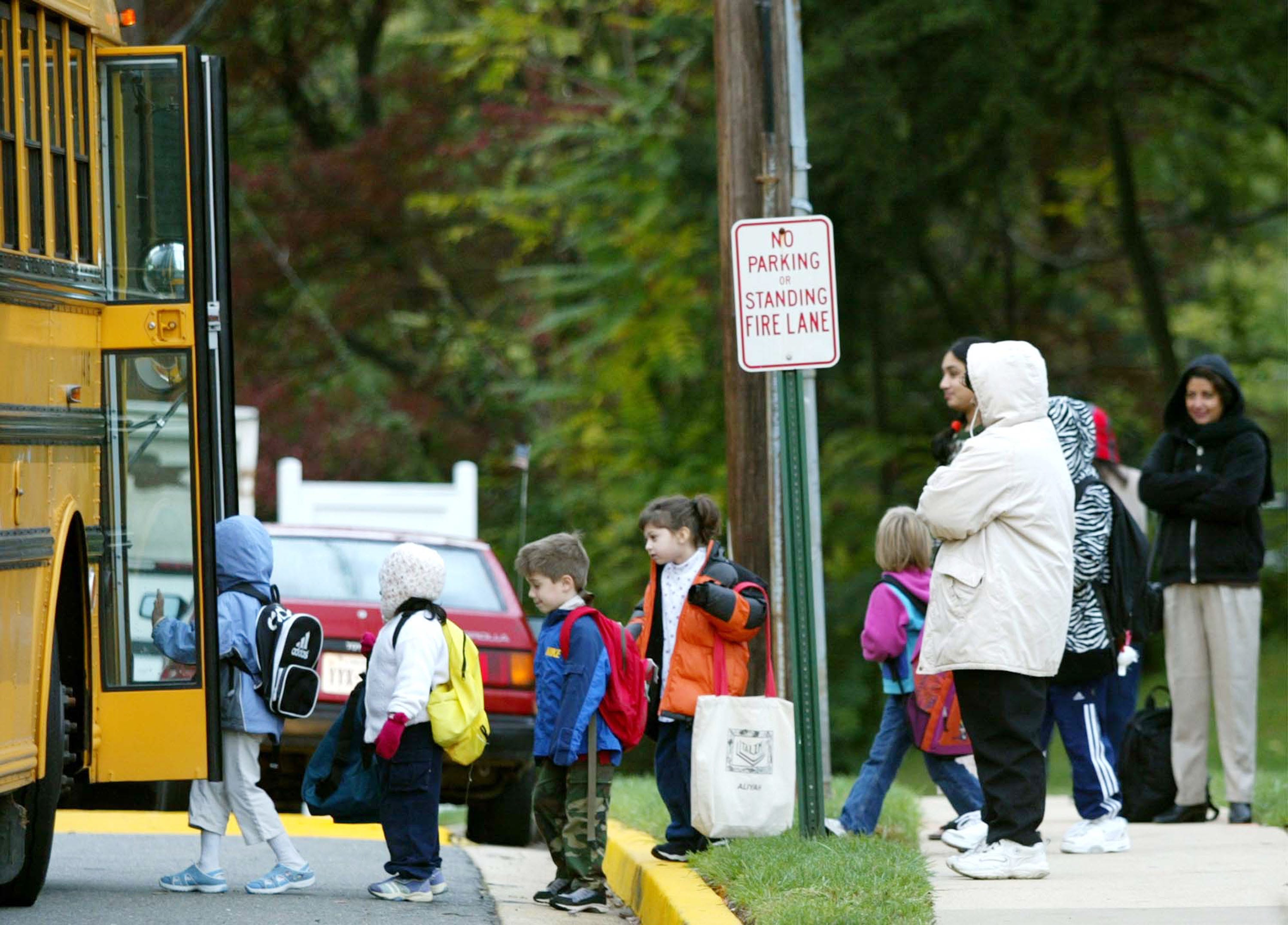 Eine Gruppe Eltern drängt sich an einer bestimmten Haltestelle zusammen, während ihre Kinder in einen Schulbus steigen.