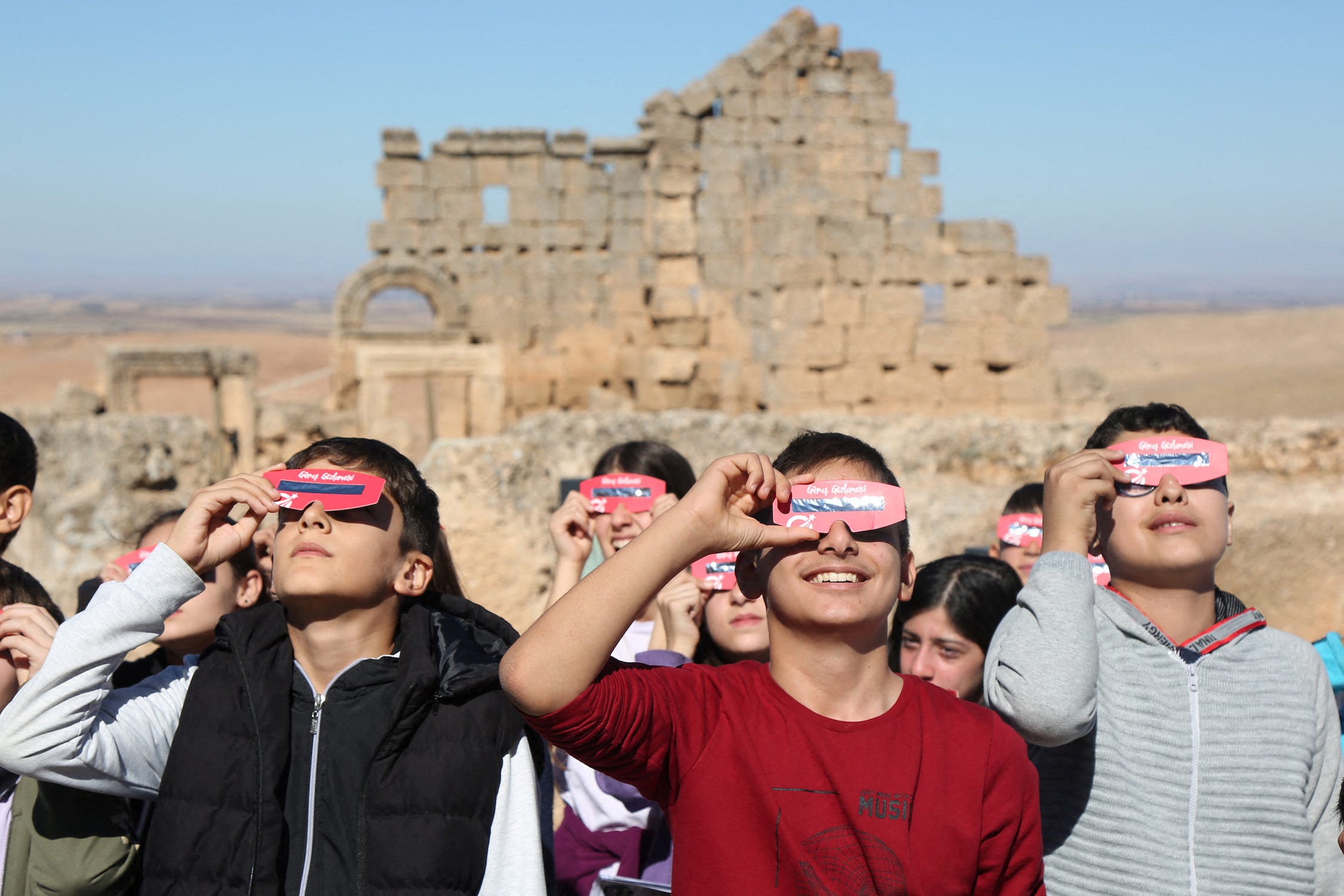 Eine Menschenmenge blickt zur Sonne auf, während sie vor einer Wand aus sandfarbenen Ziegeln in einer Wüstenlandschaft eine rosafarbene Sonnenfinsternisbrille vor die Augen hält