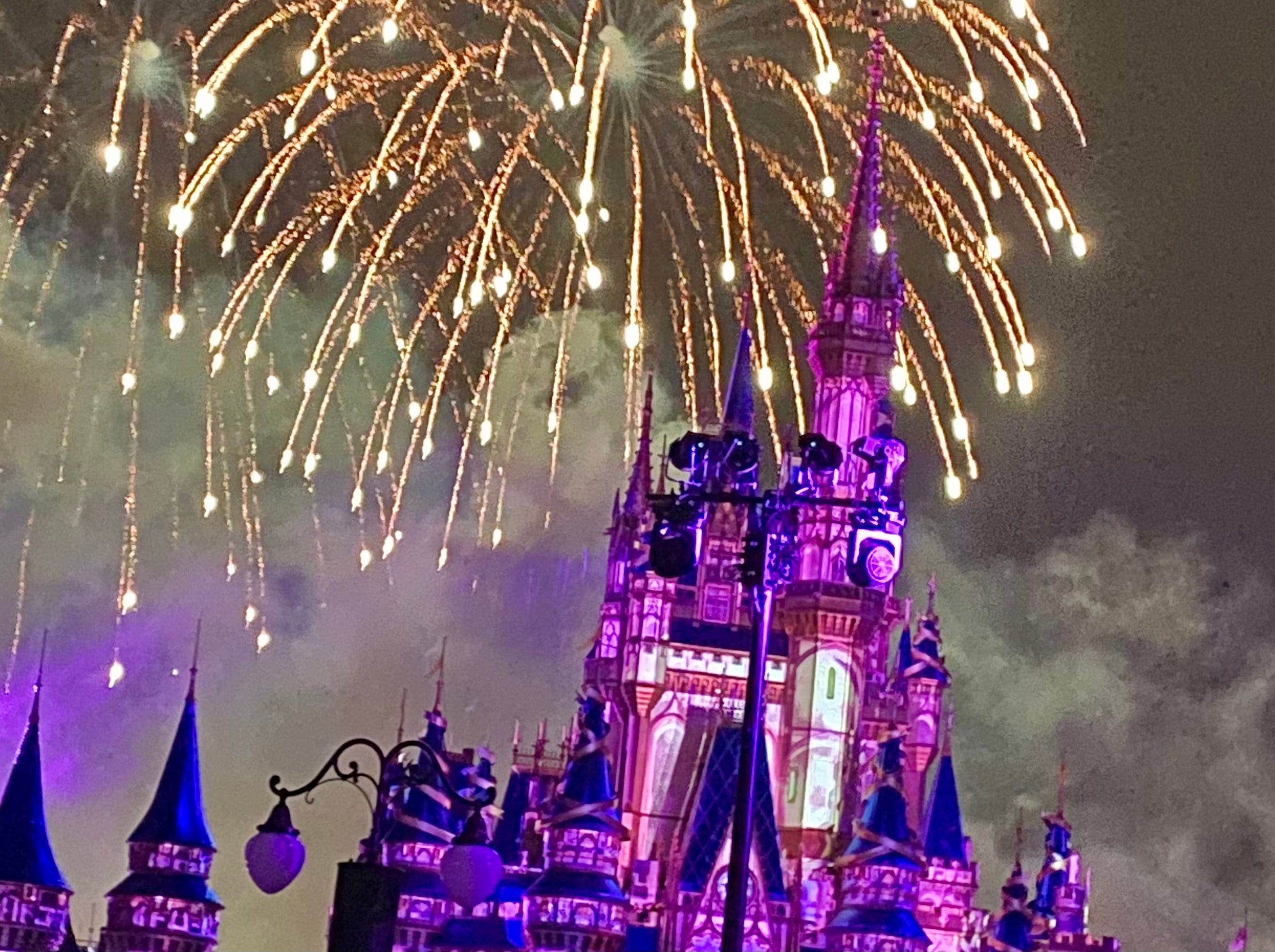 Bild eines Feuerwerks im Magic Kingdom bei Disney.  Das Schloss wird von violetten Lichtern beleuchtet und das Feuerwerk ist goldfarben.