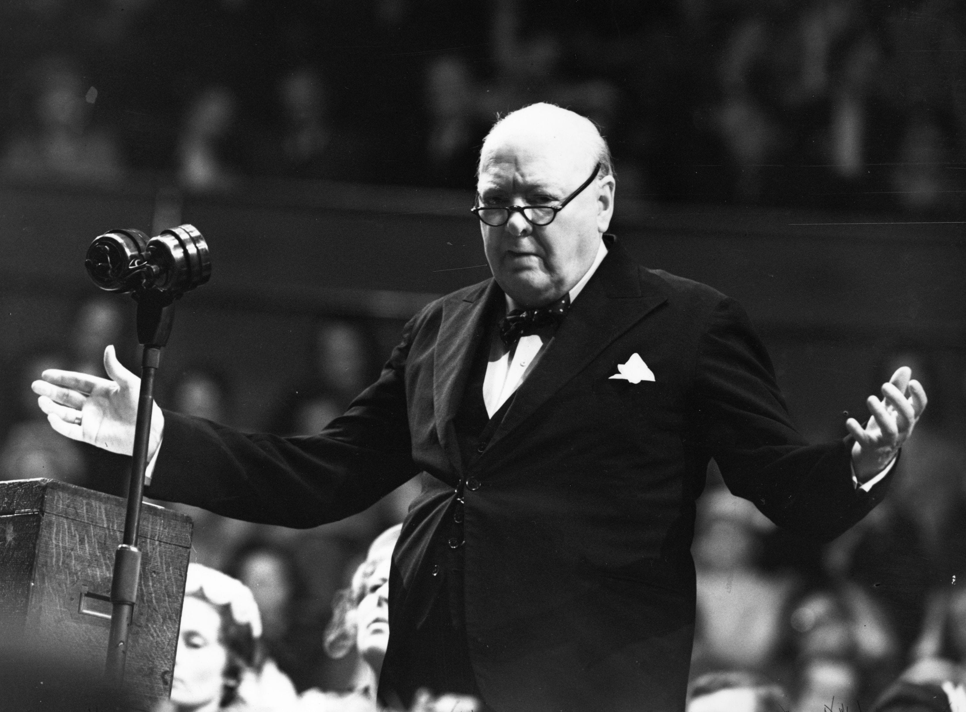 Schwarzweißfoto von Churchill im Anzug mit Fliege hinter einem Mikrofon und mit erhobenen Armen, als wollte er sagen: „Was?“