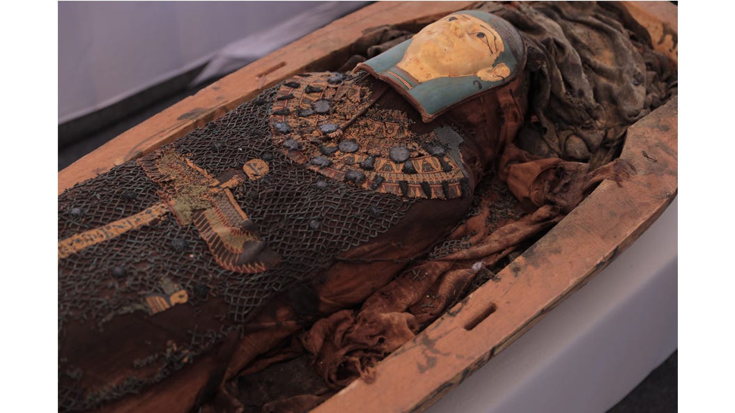 Ein Bild zeigt das Innere eines Sarkophags, in dem sich eine altägyptische Mumie in gutem Erhaltungszustand befindet, unter einem reich verzierten Karton und einer Maske, die das Gesicht des Verstorbenen darstellt.