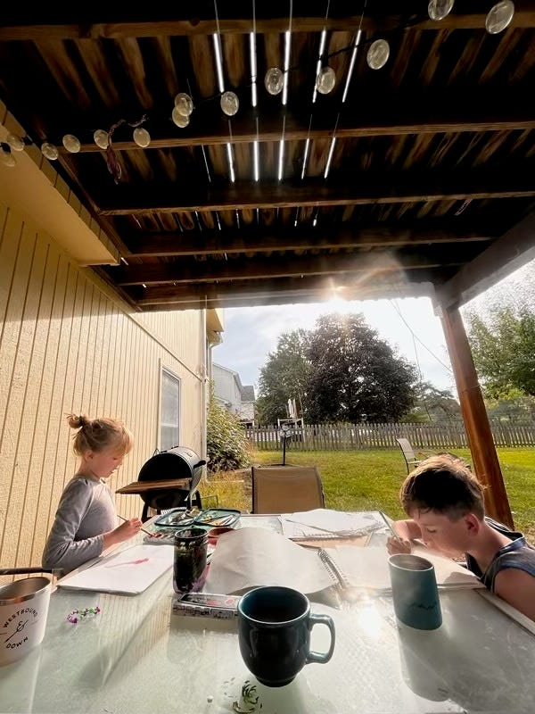 Zwei Kinder zeichnen auf einem Terrassentisch in einem grasbewachsenen Hinterhof