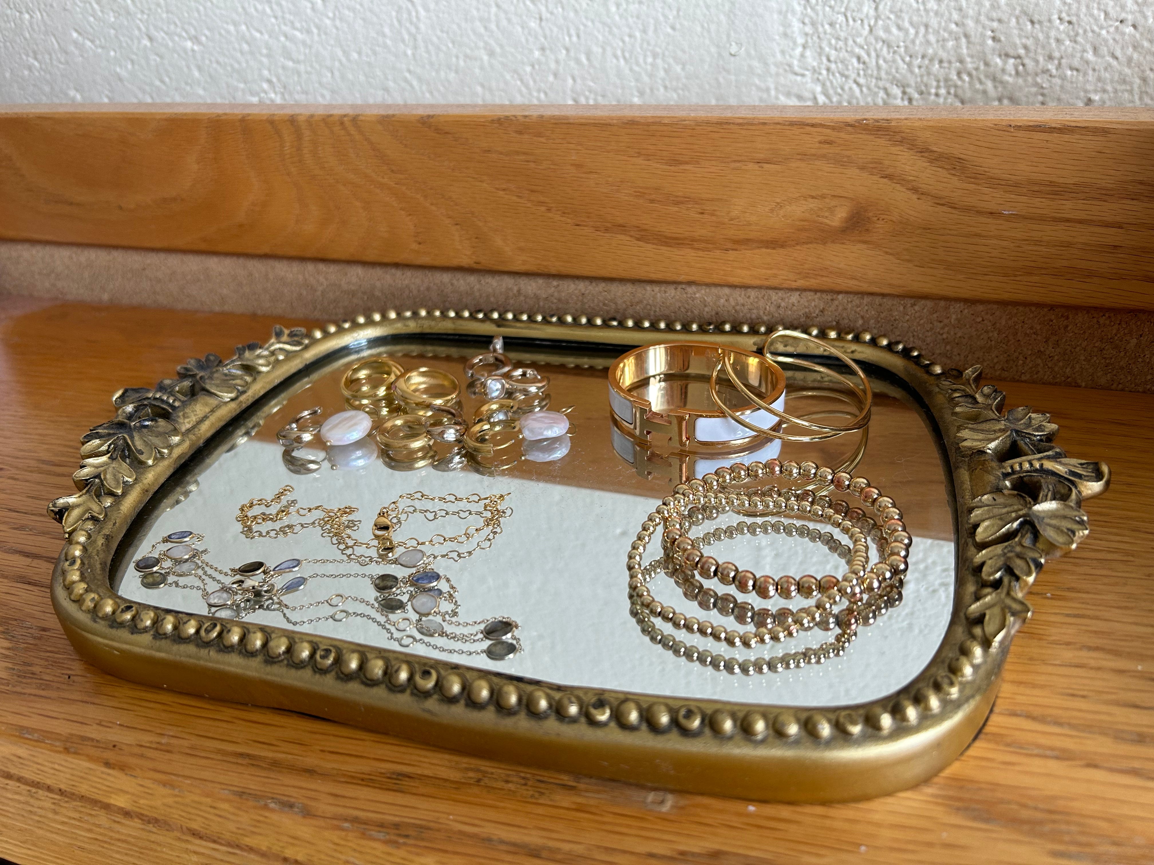 Goldenes Kosmetiktablett auf einem Holzschreibtisch.  Auf dem Tablett mit Spiegel finden goldene Armbänder und Ohrringe Platz