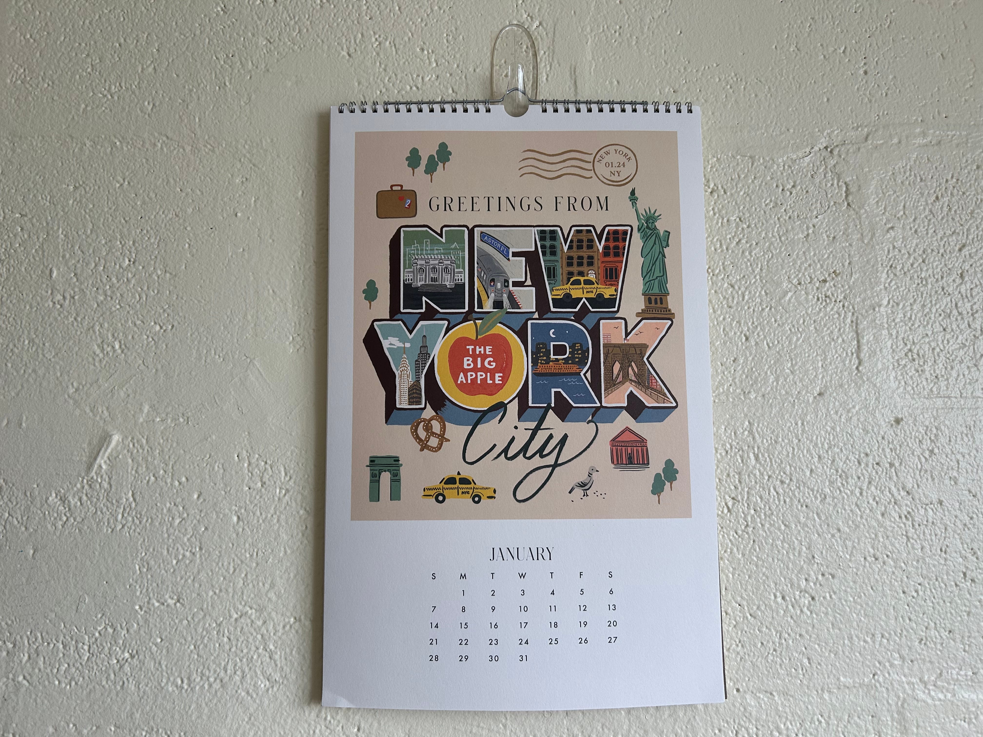 Ein Kalender auf einer weiß gestrichenen Ziegelwand.  Der Kalender hat eine rosa Grafik mit „Grüße aus New York City“ mit Illustrationen eines Taxis, der Freiheitsstatue, Tauben und einer Brezel