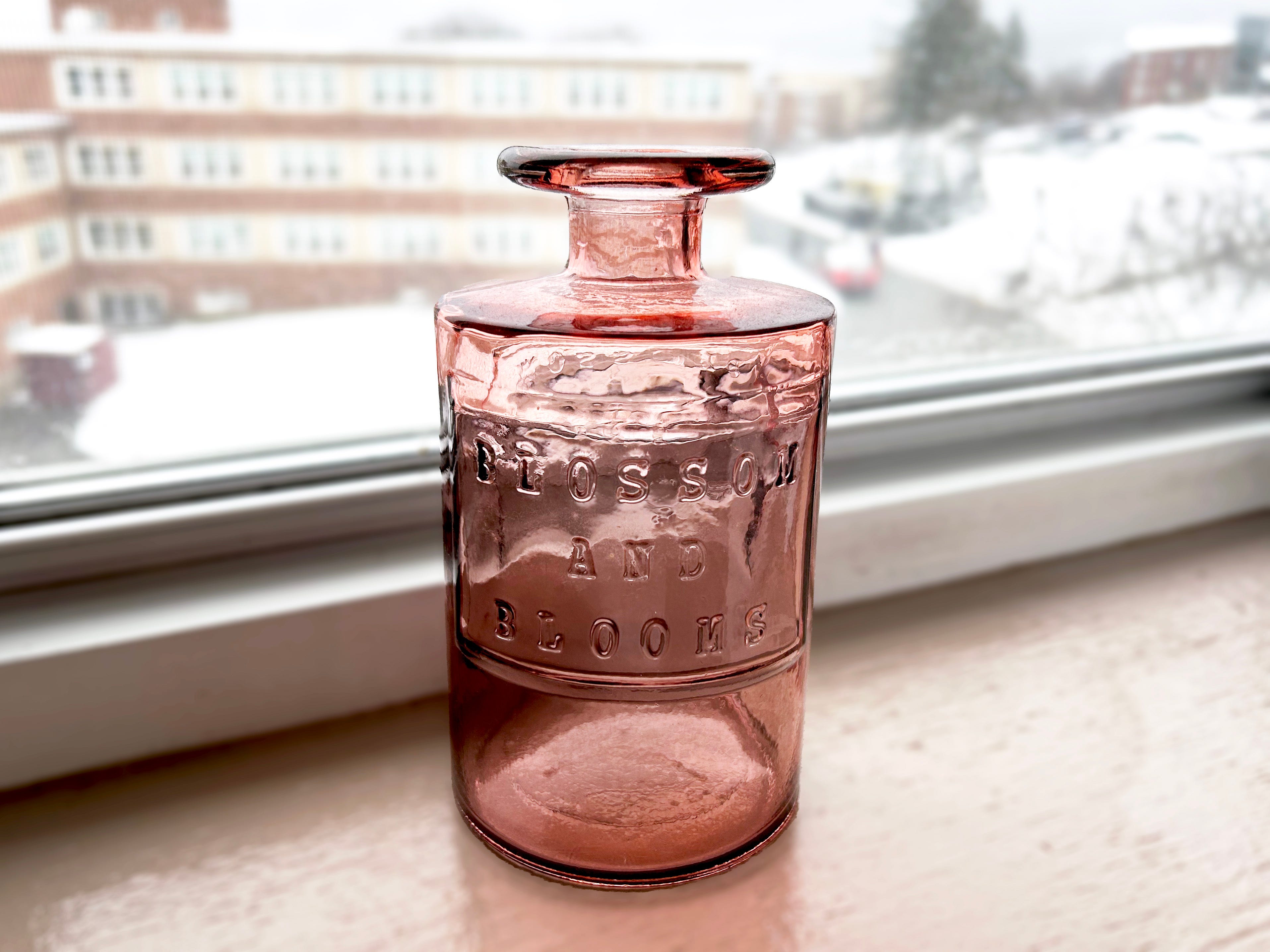 Ein rosafarbenes Glas im Apothekerstil mit der Gravur „Blüte und Blüten“.  Die Flasche steht auf einem Holzsims neben einem Fenster
