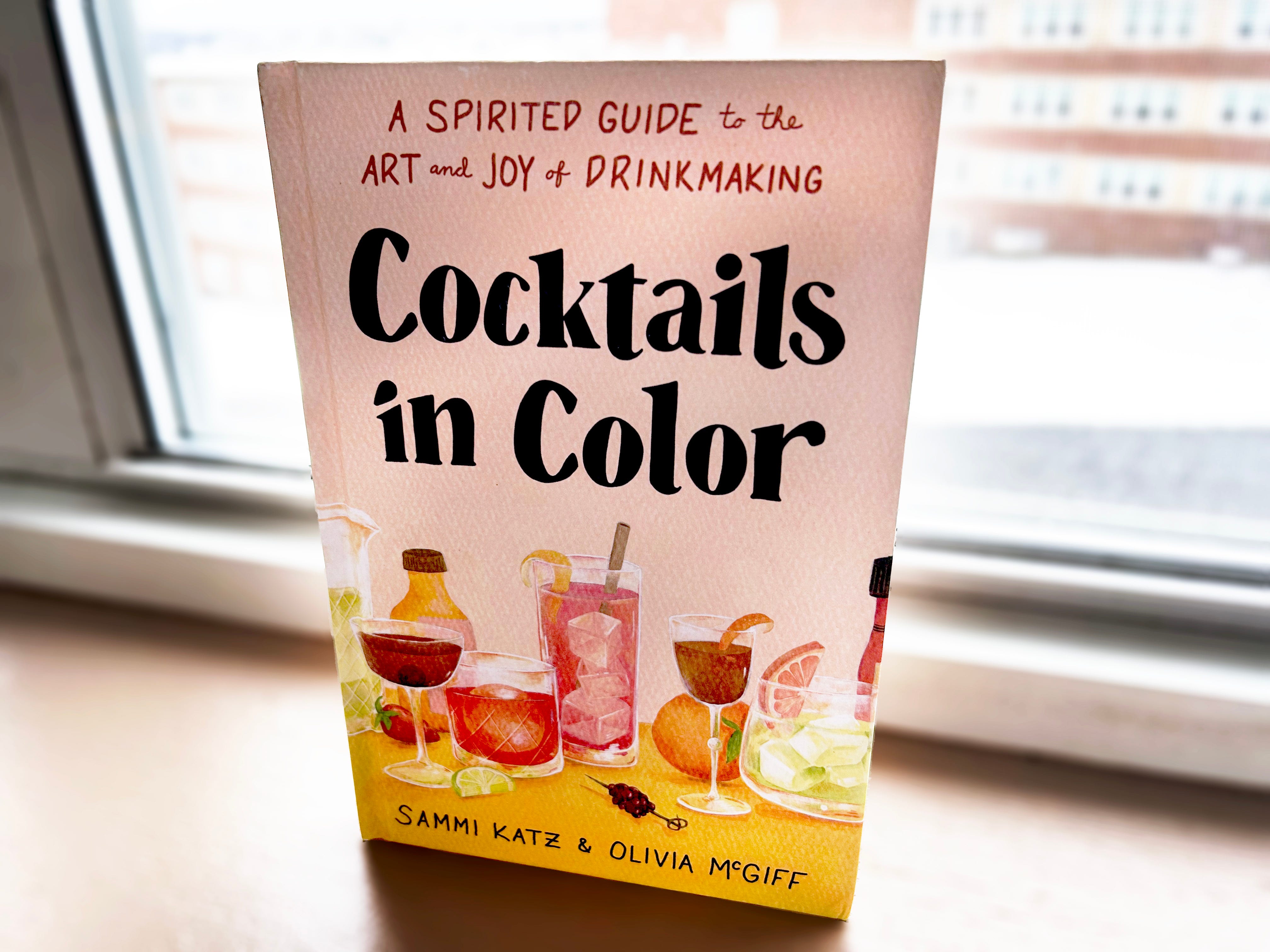 Auf einem Holzbrett neben einem Fenster liegt ein Buch mit dem Titel „Cocktails in Colour“ mit rosa Hintergrund und Abbildungen bunter Cocktails