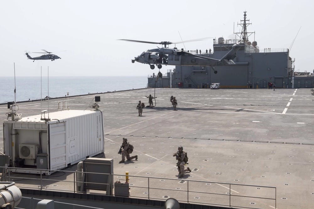 Marineinfanteristen stehen auf dem Flugdeck eines ESB, während darüber ein Hubschrauber schwebt