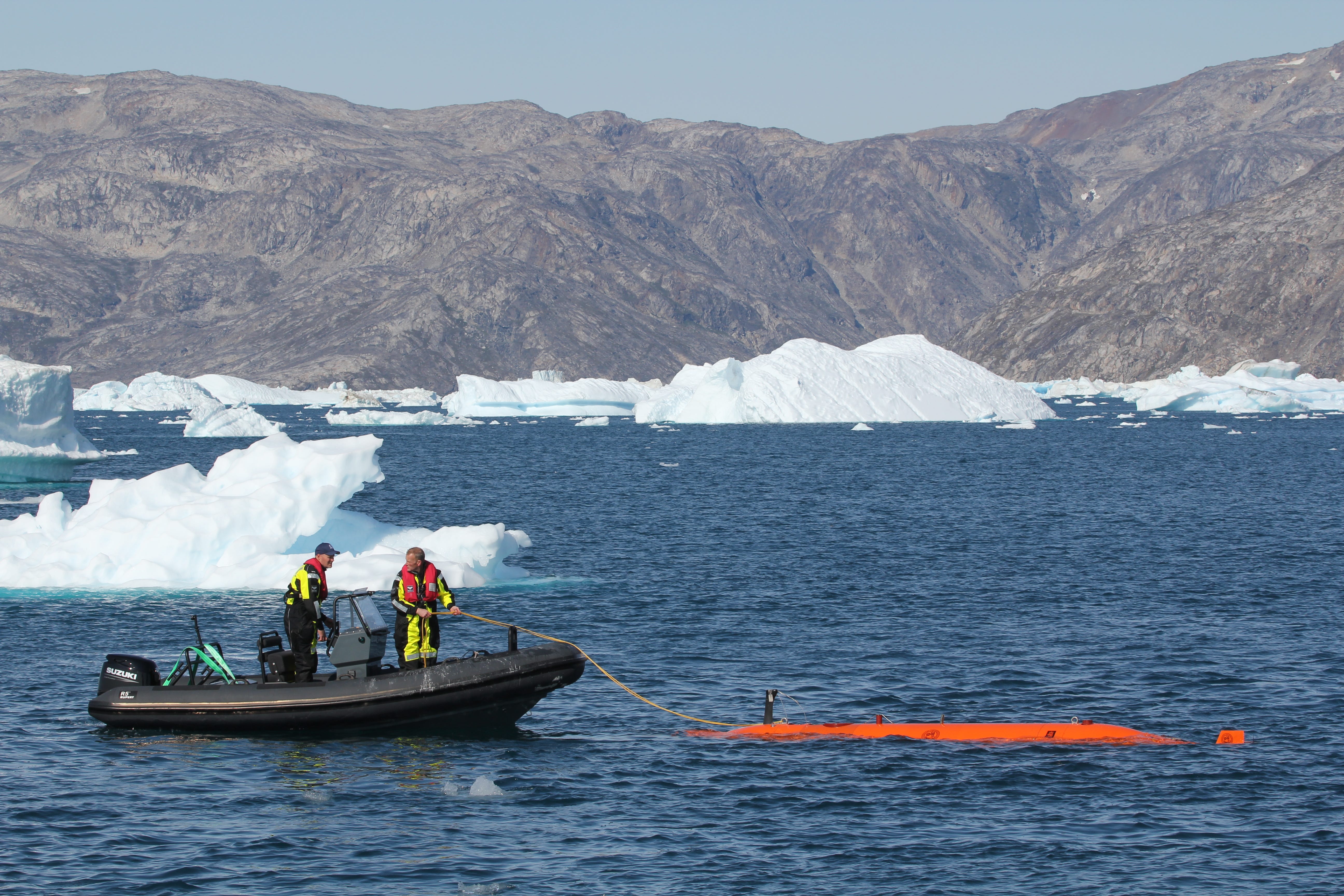 Zwei Personen auf einem Schlauchboot im Meer, ein Seil haltend, das an das Ran-Tauchboot gebunden ist, mit Eis und Bergen im Hintergrund