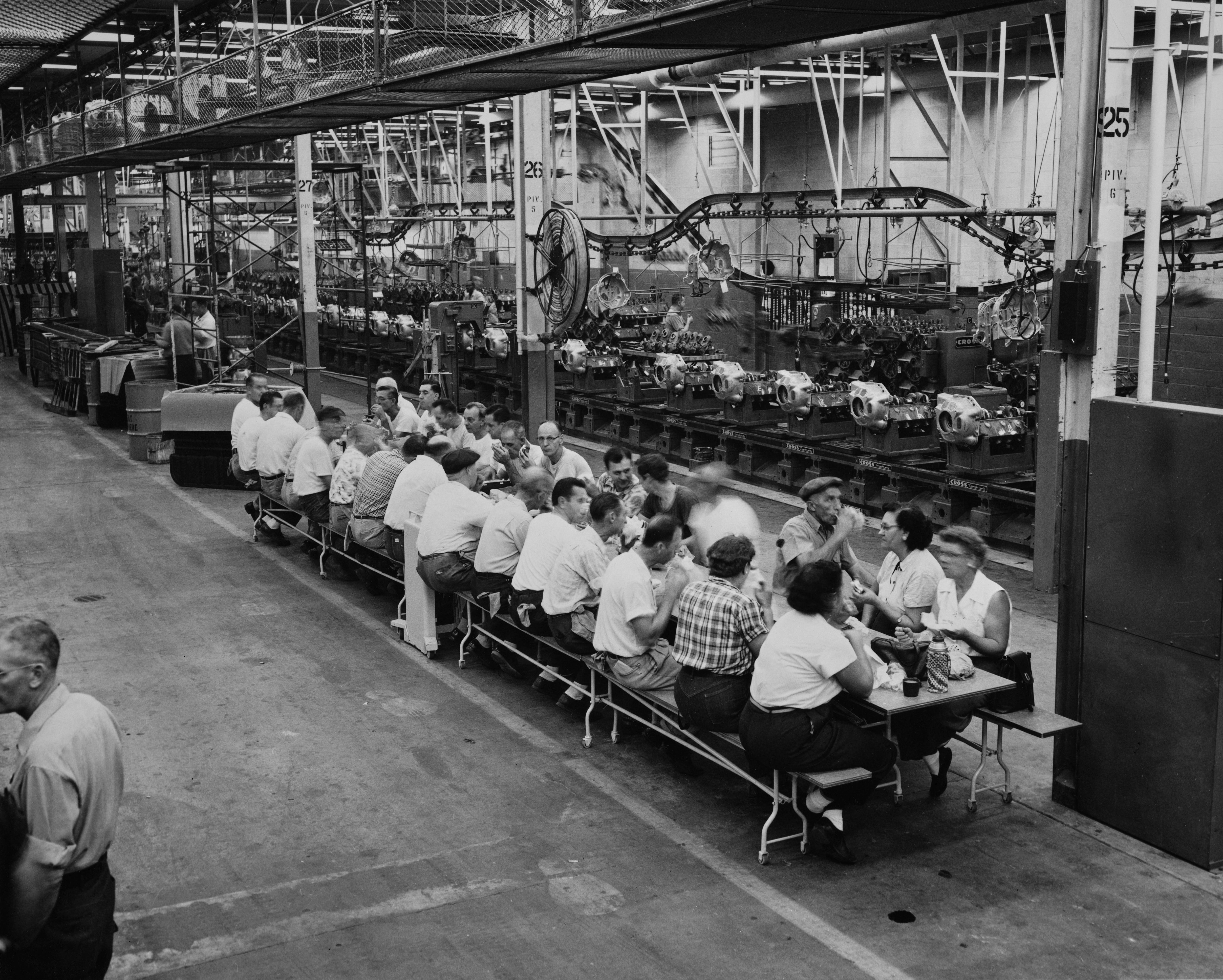 Eine Gruppe von Arbeitern sitzt auf einer langen Bank beim Mittagessen in der Chrysler Mound Road Engine-Fabrik in Detroit, Michigan, um 1955.