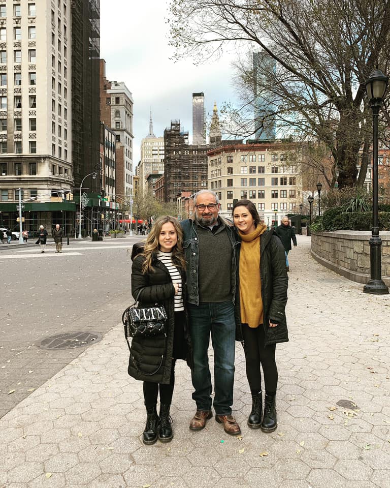 Rachel Volk steht neben ihrem Vater und ihrer Schwester auf einer Straße in New York City.  Dahinter sind Gebäude zu erkennen.