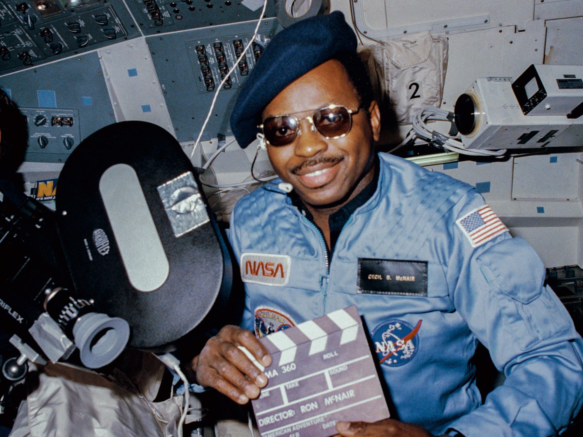 Der Astronaut Ron McNair trägt eine Sonnenbrille und eine Baskenmütze und hält eine dünne Schiefertafel in einem Raumschiff